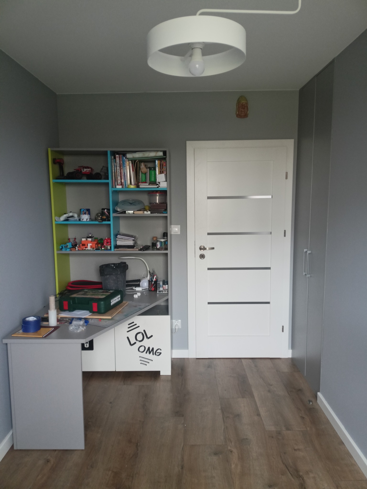 Szary pokój dziecięcy po malowaniu: biurko z regałem, białe drzwi z poziomymi paskami, drewniana podłoga. Na ścianie dekoracja w kształcie głowy Buddy.