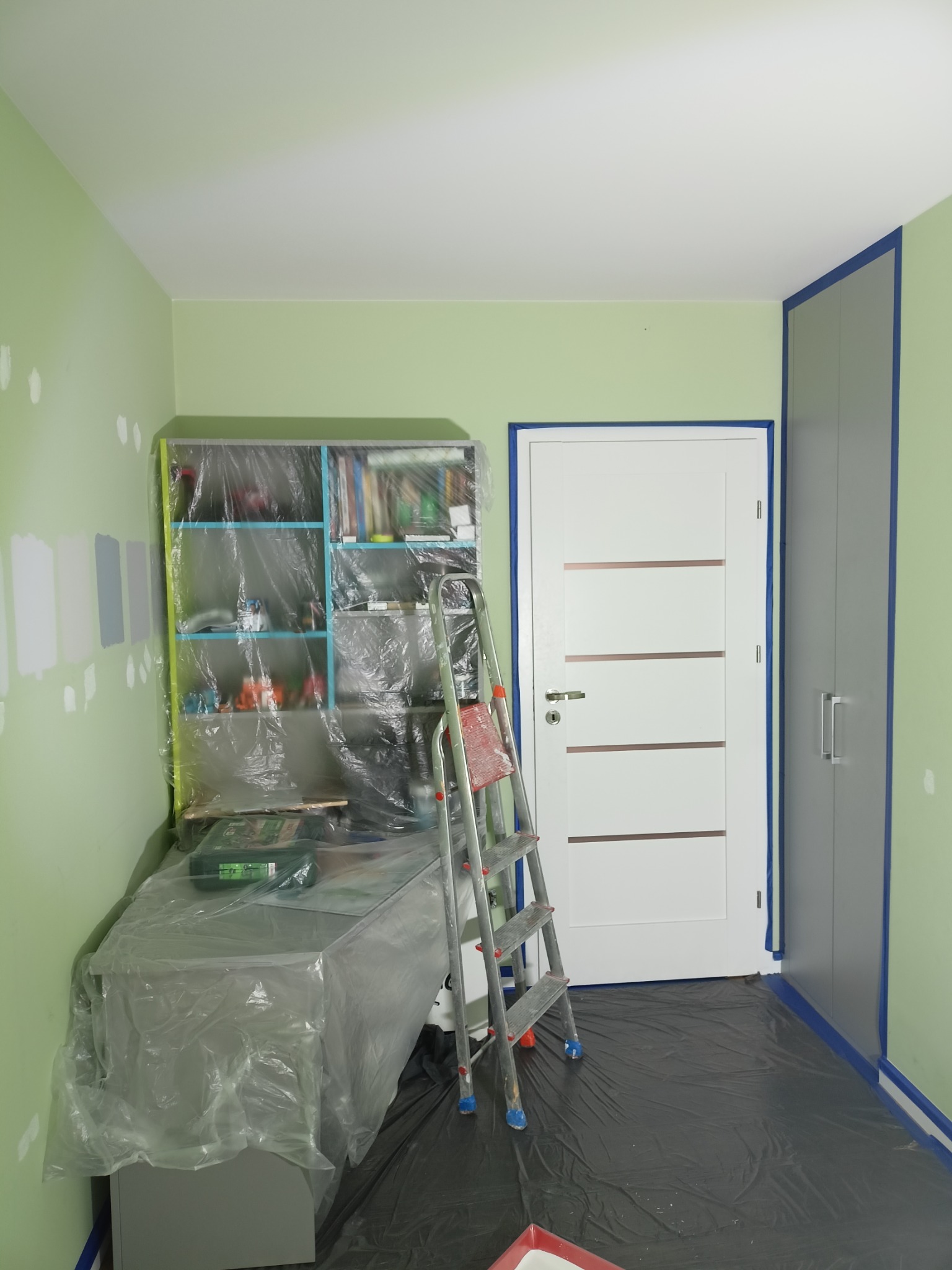 Pokój w trakcie malowania: zabezpieczone meble folią, drabina oparta o ścianę, testowe kolory na ścianie, taśma malarska przy drzwiach i szafie.