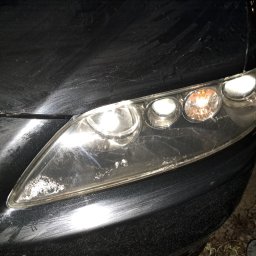 Drobne naprawy pojazdów, konserwacja ,sprzątanie Adam Wołoszyn - Brudny, czarny samochód z bliska, z widocznymi zarysowaniami i osadem na lakierze oraz matową lampą przednią z zapalonymi światłami.