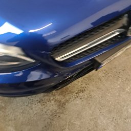 Drobne naprawy pojazdów, konserwacja ,sprzątanie Adam Wołoszyn - Fragment niebieskiego samochodu, widoczny reflektor, grill i część maski. Na podłodze stoi but. Ujęcie z bliska, z perspektywy osoby stojącej obok pojazdu.