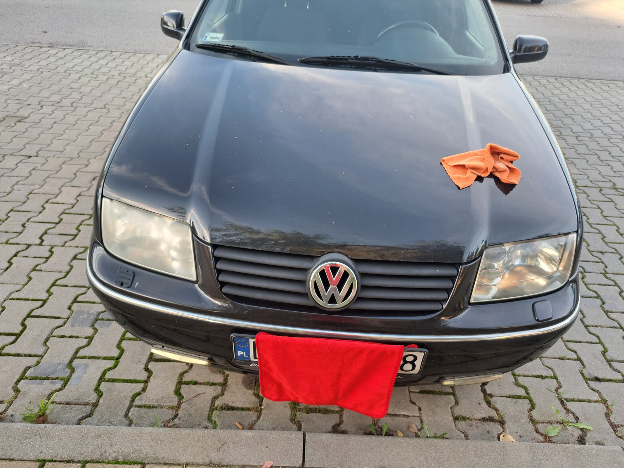 Czarny samochód Volkswagen z pomarańczową ściereczką na masce i czerwoną na tablicy rejestracyjnej, zaparkowany na chodniku z kostki.