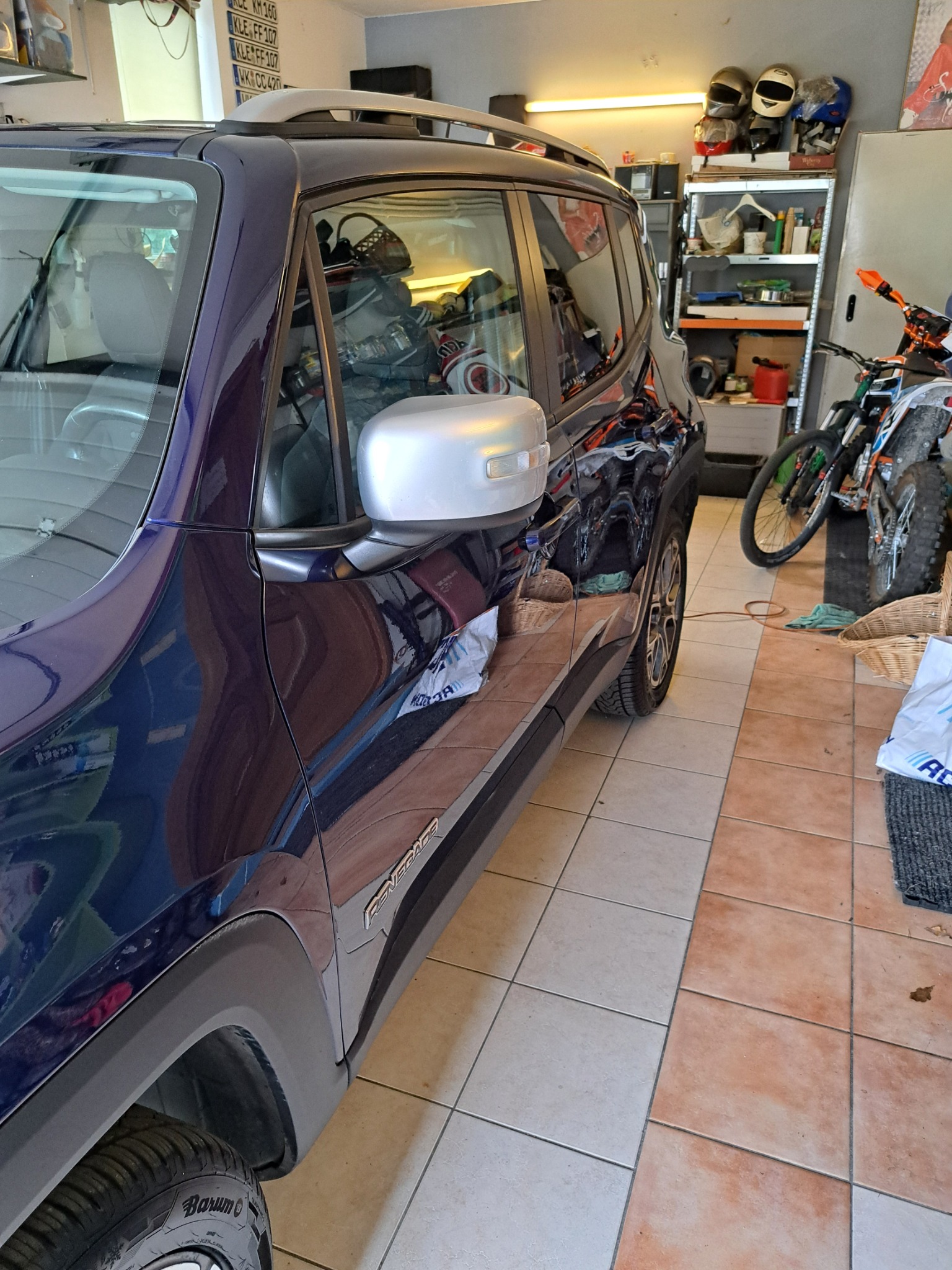 Błyszczący, granatowy Jeep Renegade zaparkowany w garażu, widoczne detale karoserii i opony Barum, w tle półki z kaskami motocyklowymi i motocykl crossowy.