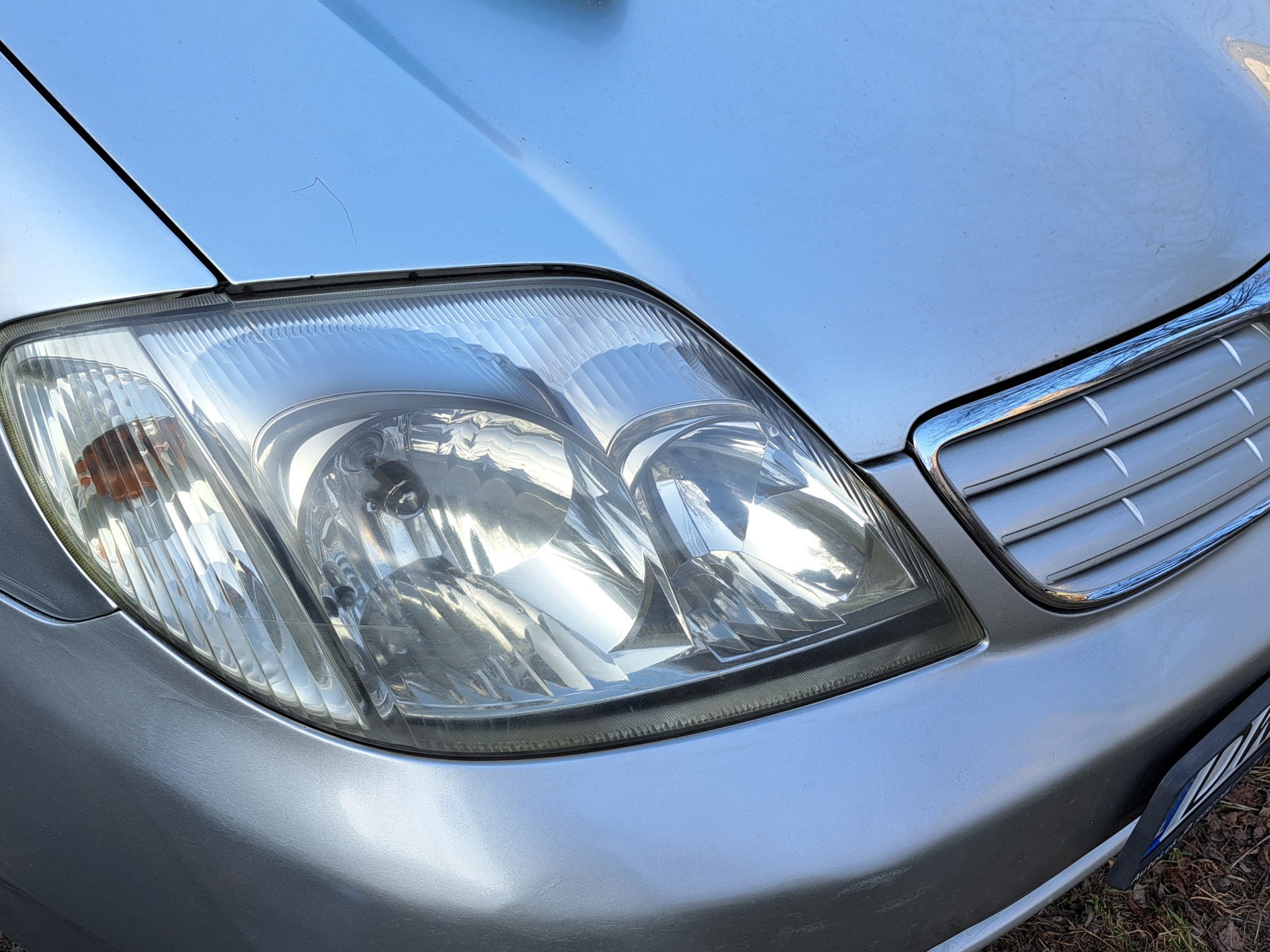 Błyszczący reflektor przedni srebrnego samochodu z widocznymi drobnymi zarysowaniami na karoserii i chromowanym grillem w słoneczny dzień.