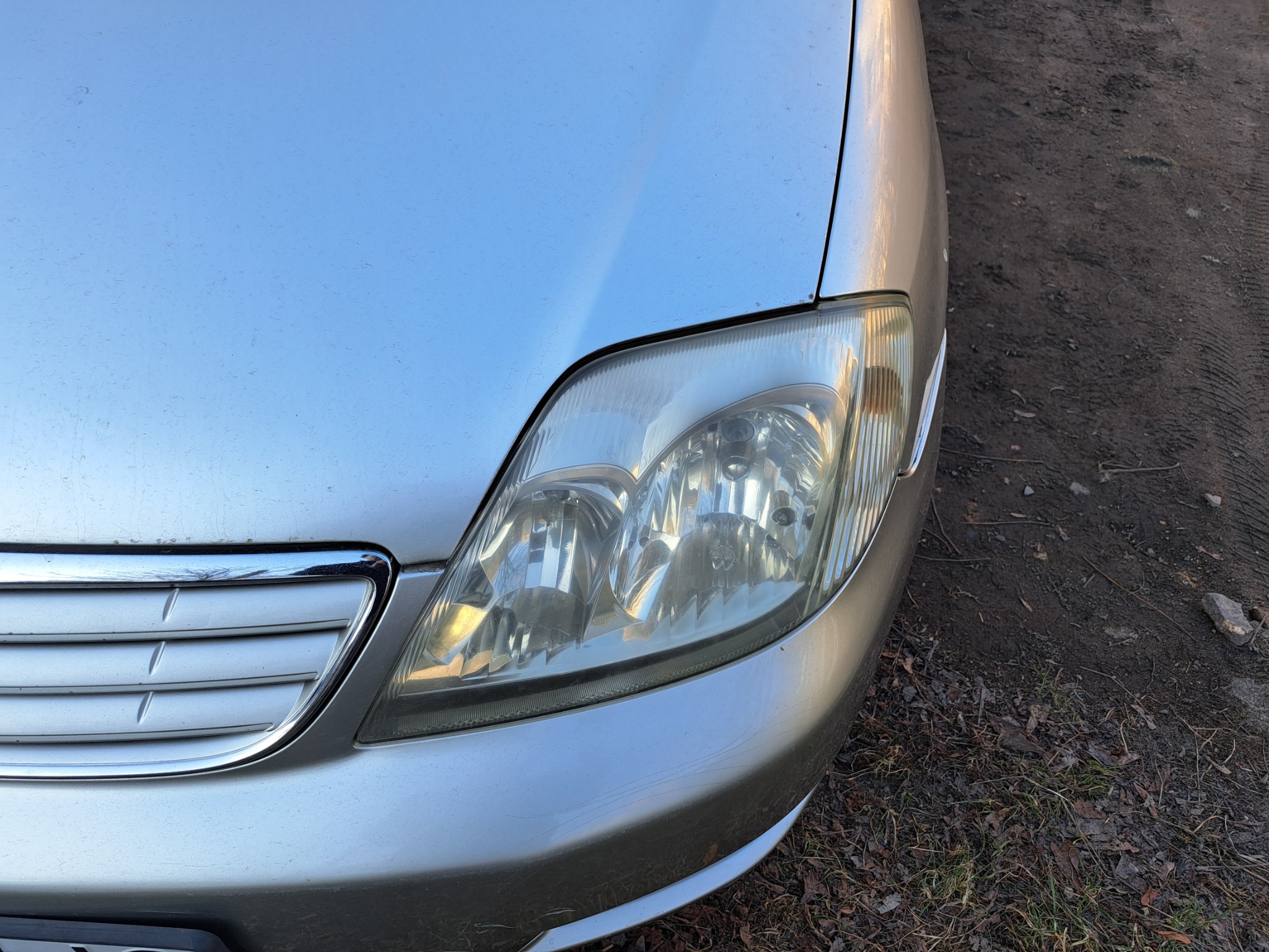 Przedni fragment srebrnego samochodu z widoczną lampą i grillem, zarysowaniami na zderzaku i brudnym tłem.