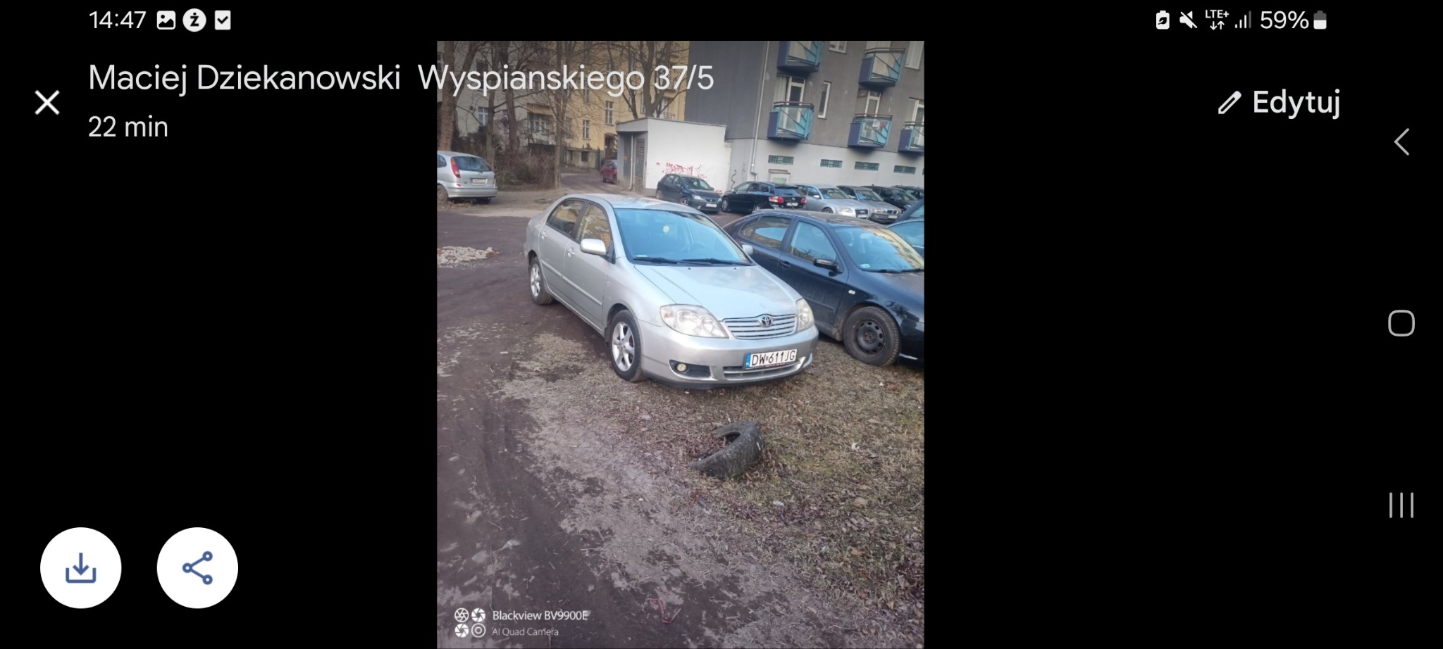 Srebrny samochód Toyota Corolla zaparkowany na parkingu w pobliżu budynku mieszkalnego, widoczna opona leżąca na ziemi.