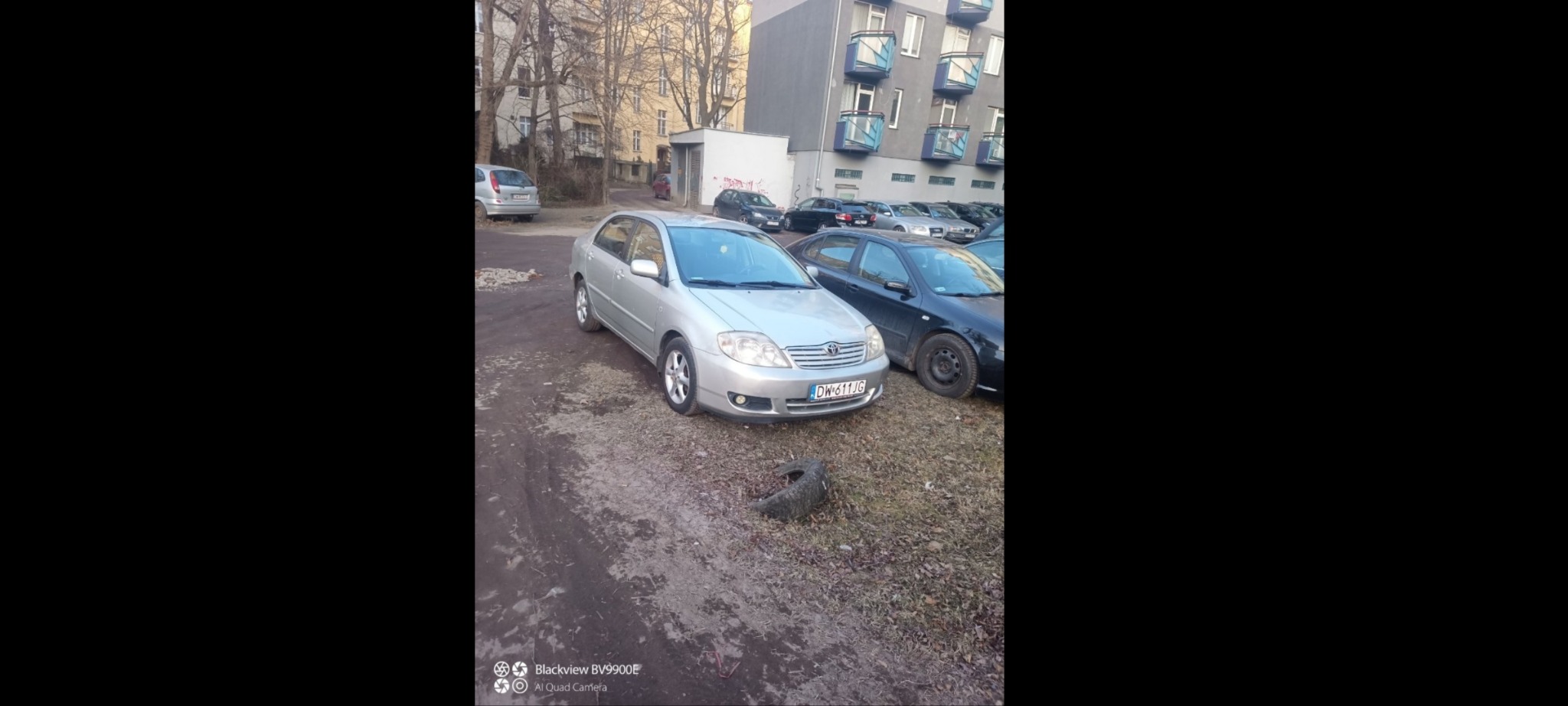 Srebrny samochód Toyota Corolla zaparkowany na placu z widocznym starym oponą na ziemi, w tle budynek mieszkalny z balkonami.