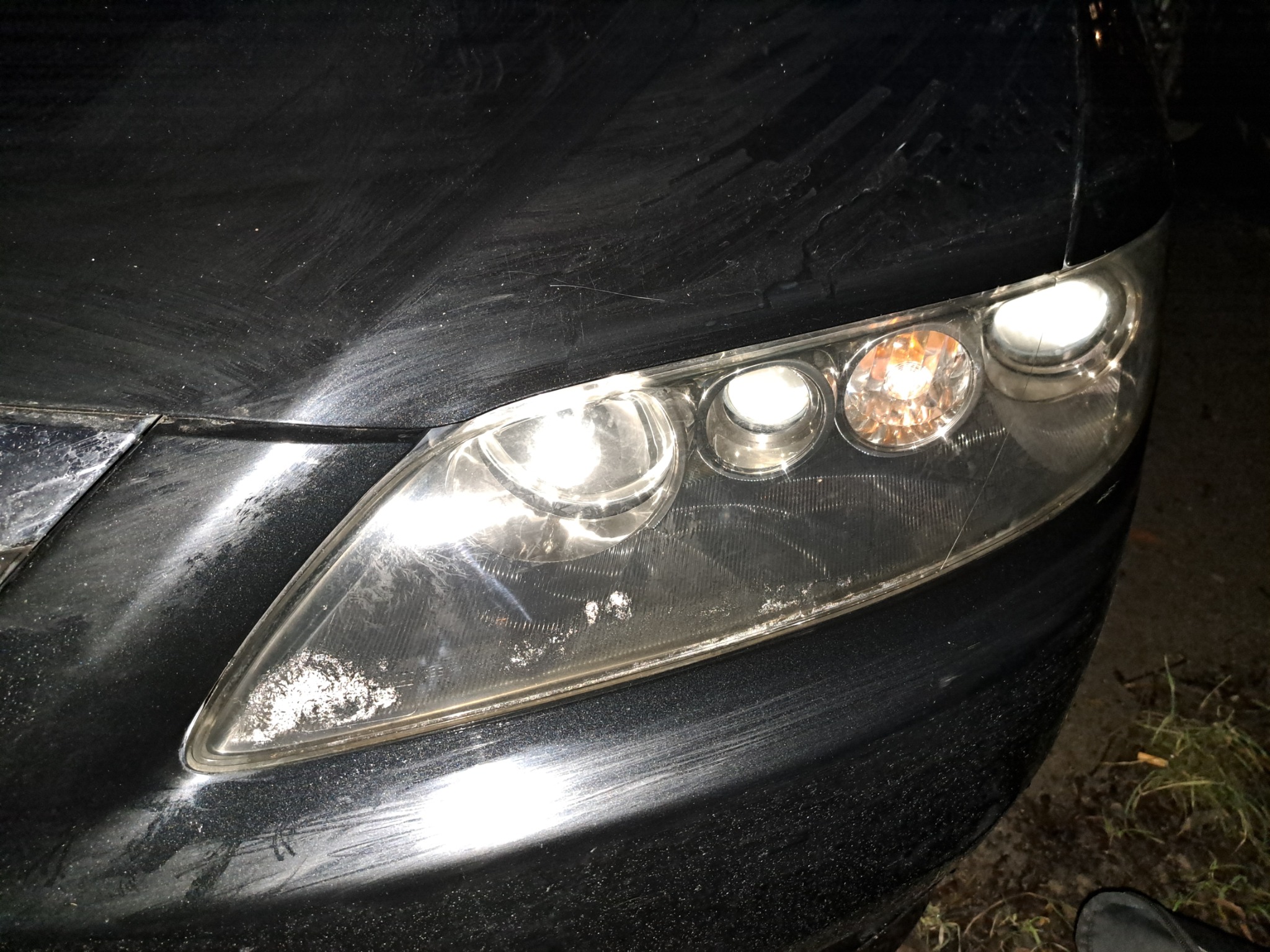 Brudny, czarny samochód z bliska, z widocznymi zarysowaniami i osadem na lakierze oraz matową lampą przednią z zapalonymi światłami.