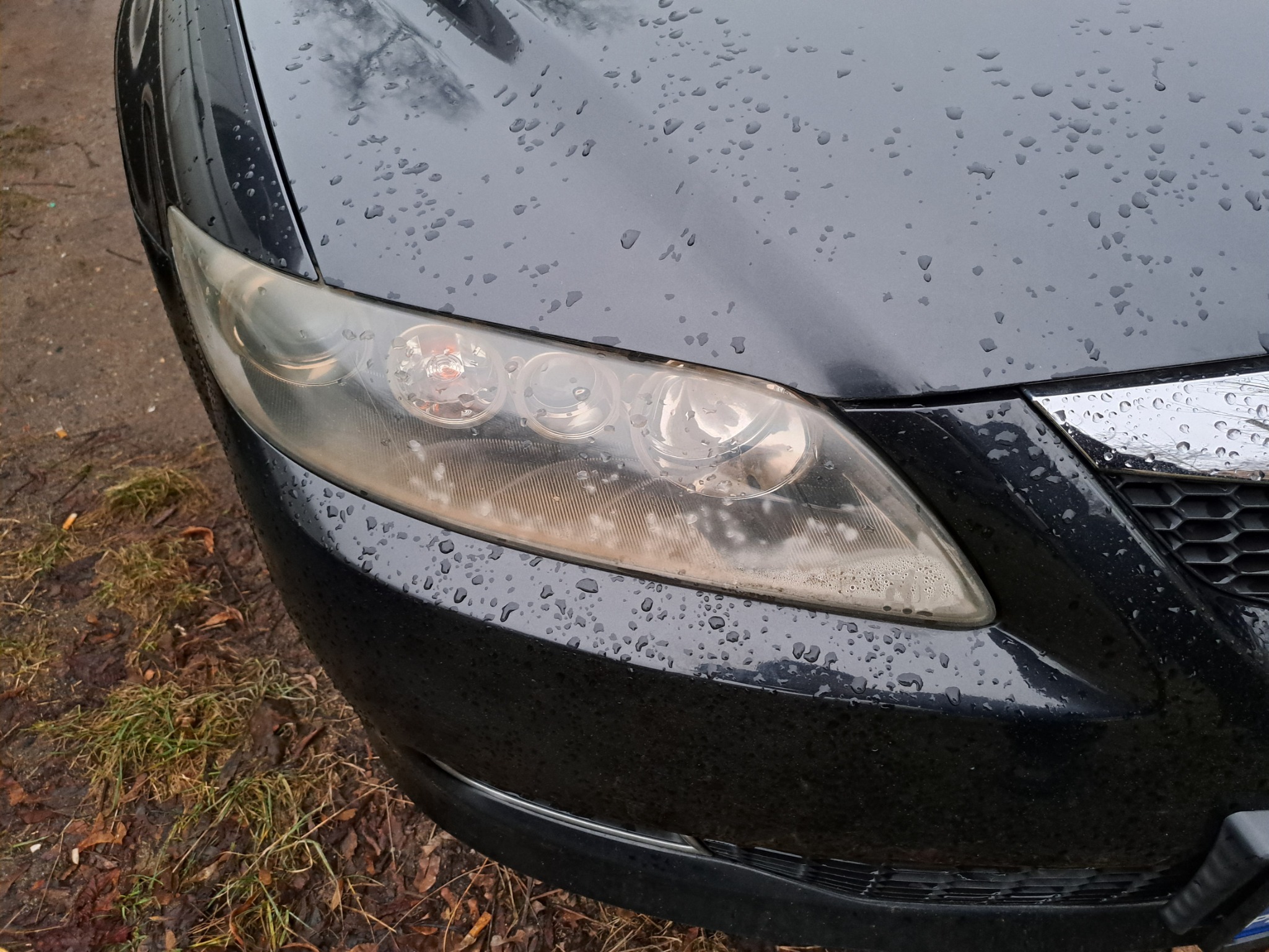 Przedni reflektor samochodu z widocznymi śladami zużycia i kroplami wody na karoserii, sfotografowany w pochmurny dzień.