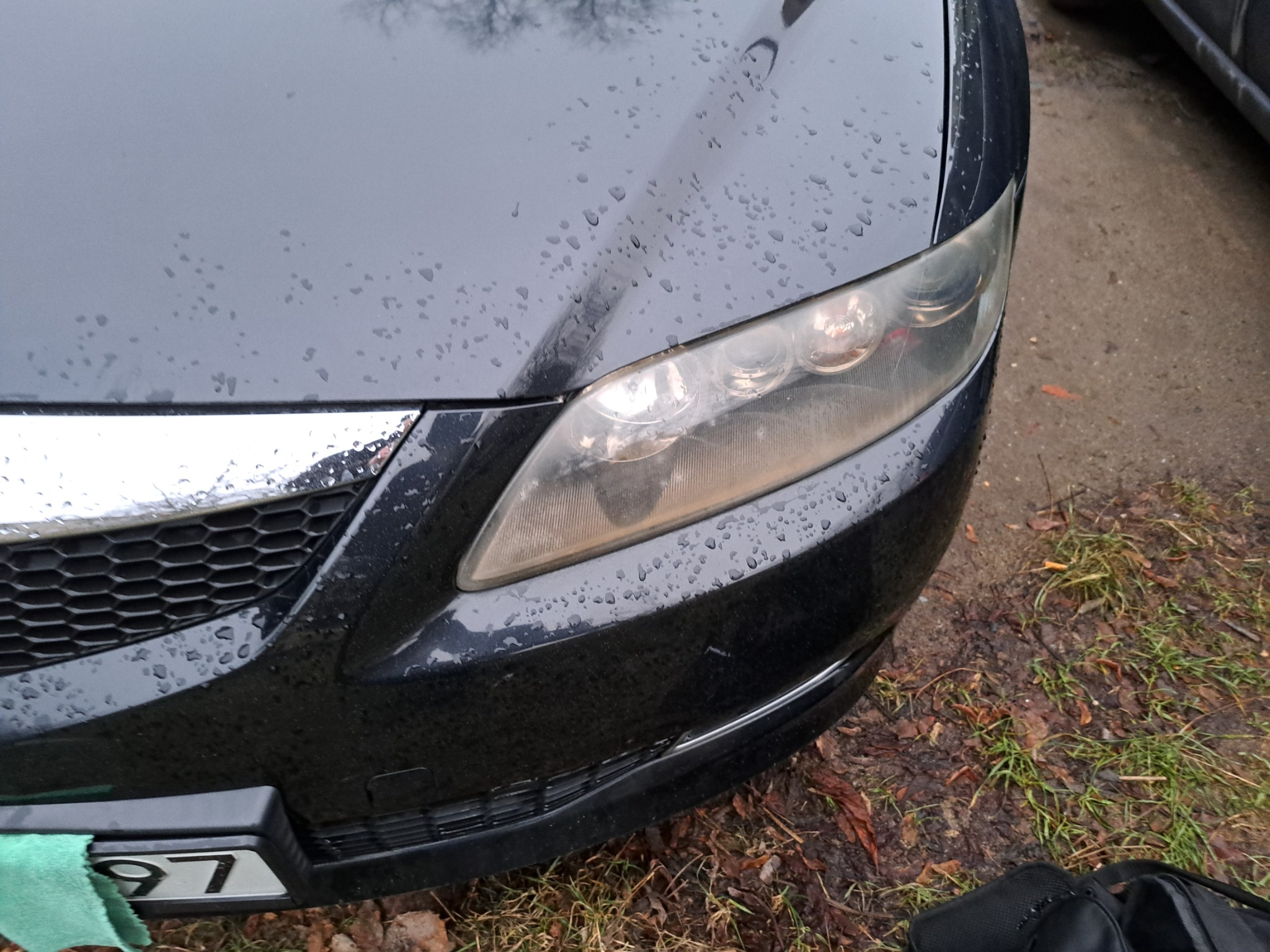 Przednia część czarnego samochodu we Wrocławiu, pokryta kroplami wody po deszczu, z widocznym reflektorem wymagającym polerowania i fragmentem tablicy rejestracyjnej.