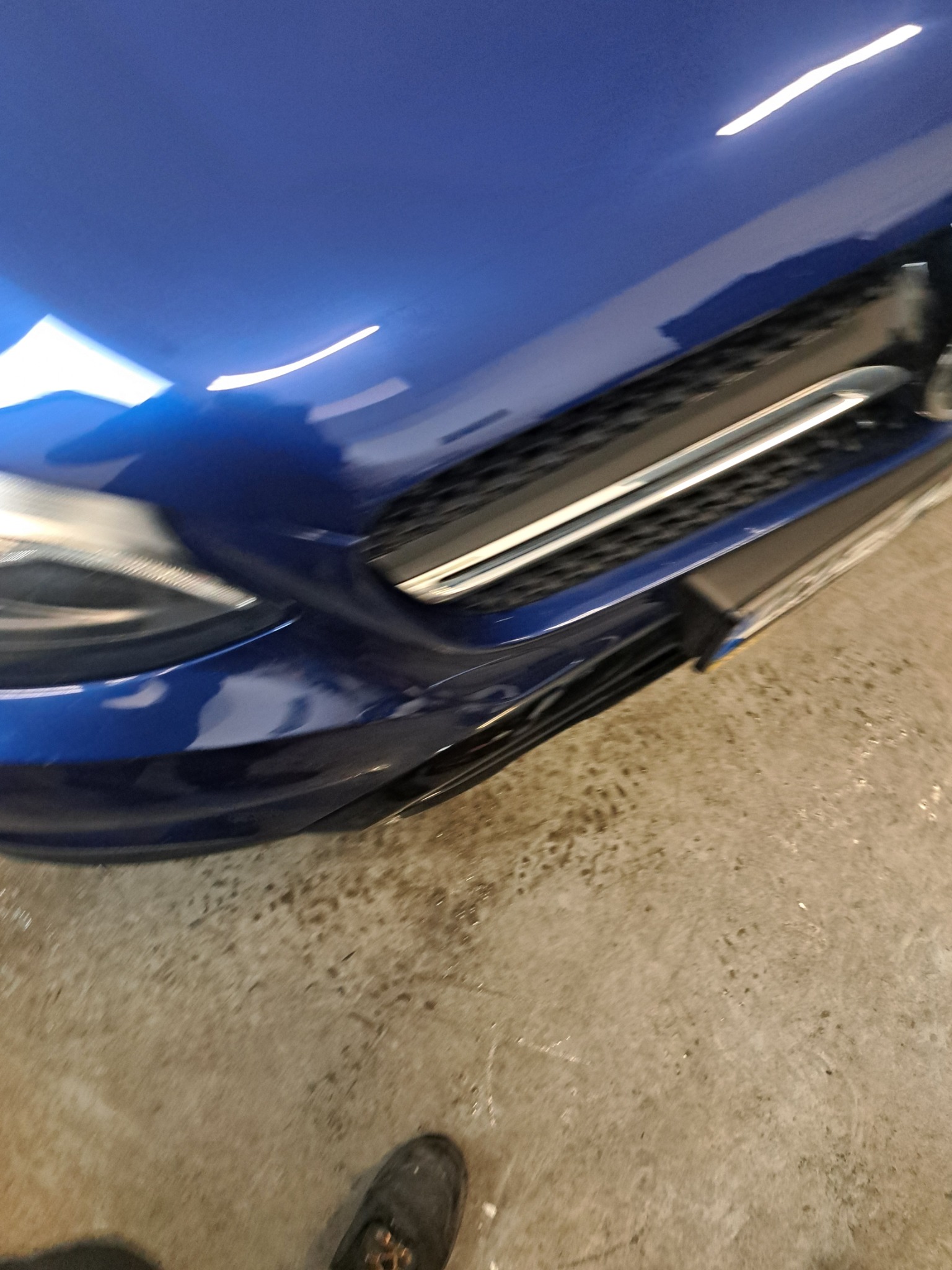 Fragment niebieskiego samochodu, widoczny reflektor, grill i część maski. Na podłodze stoi but. Ujęcie z bliska, z perspektywy osoby stojącej obok pojazdu.