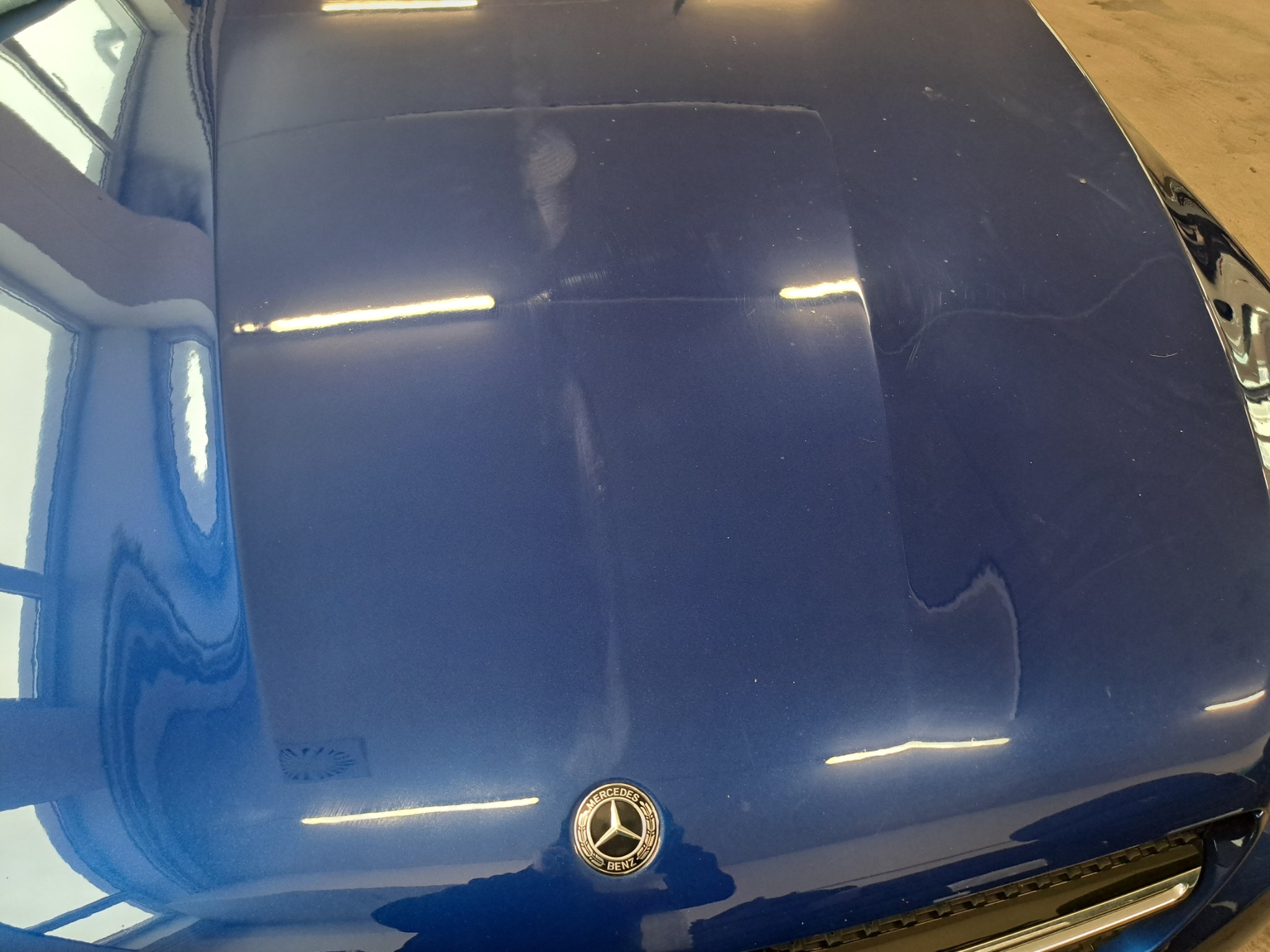 Błękitny lakier maski Mercedesa przed polerowaniem we Wrocławiu. Widoczne zarysowania i hologramy pod światło. Logo producenta w dolnej części kadru.