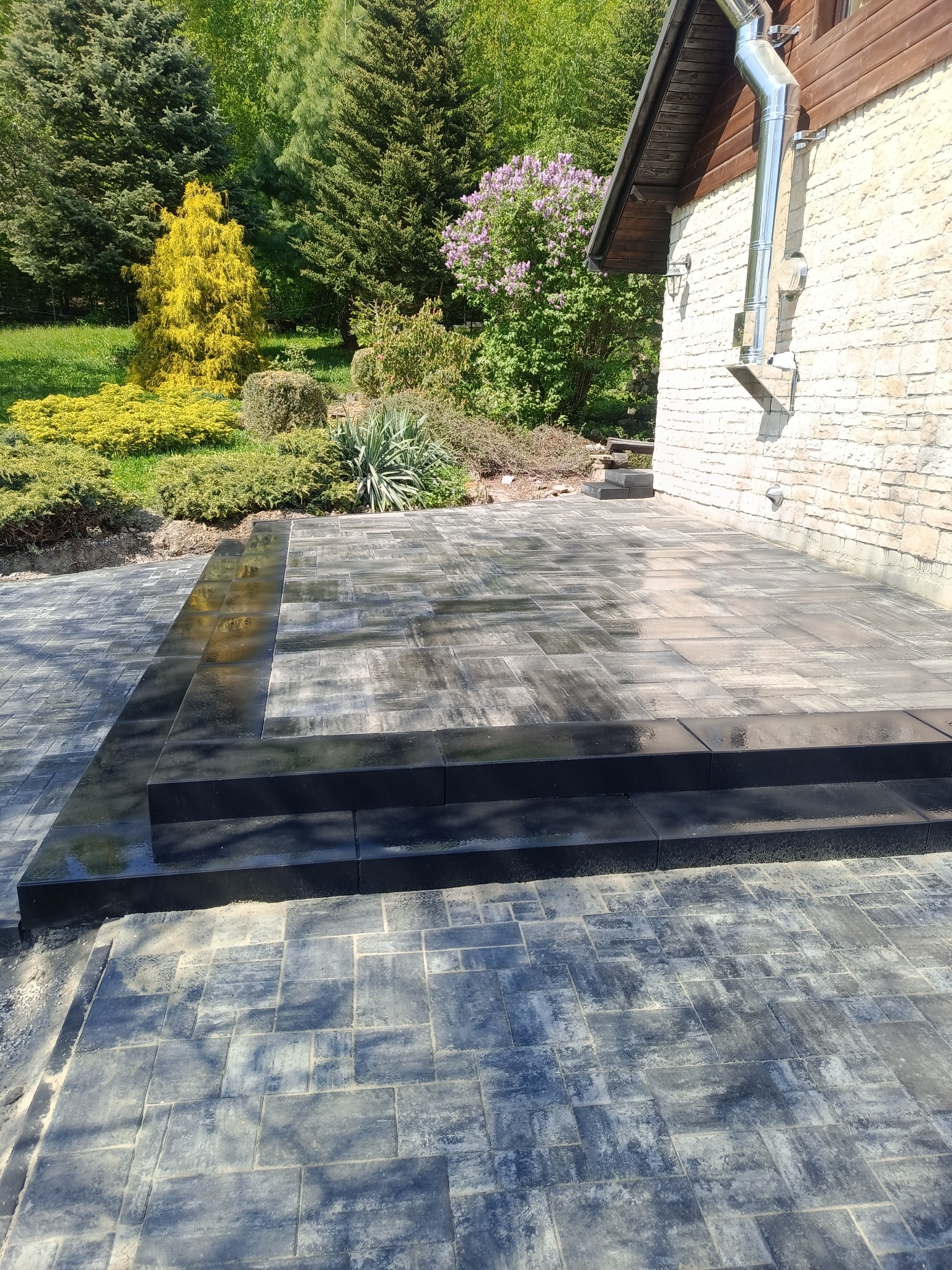 Ułożona kostka brukowa w odcieniach szarości tworząca taras przy domu z kamienną elewacją. Widoczne schody z czarnej kostki, zieleń w tle.