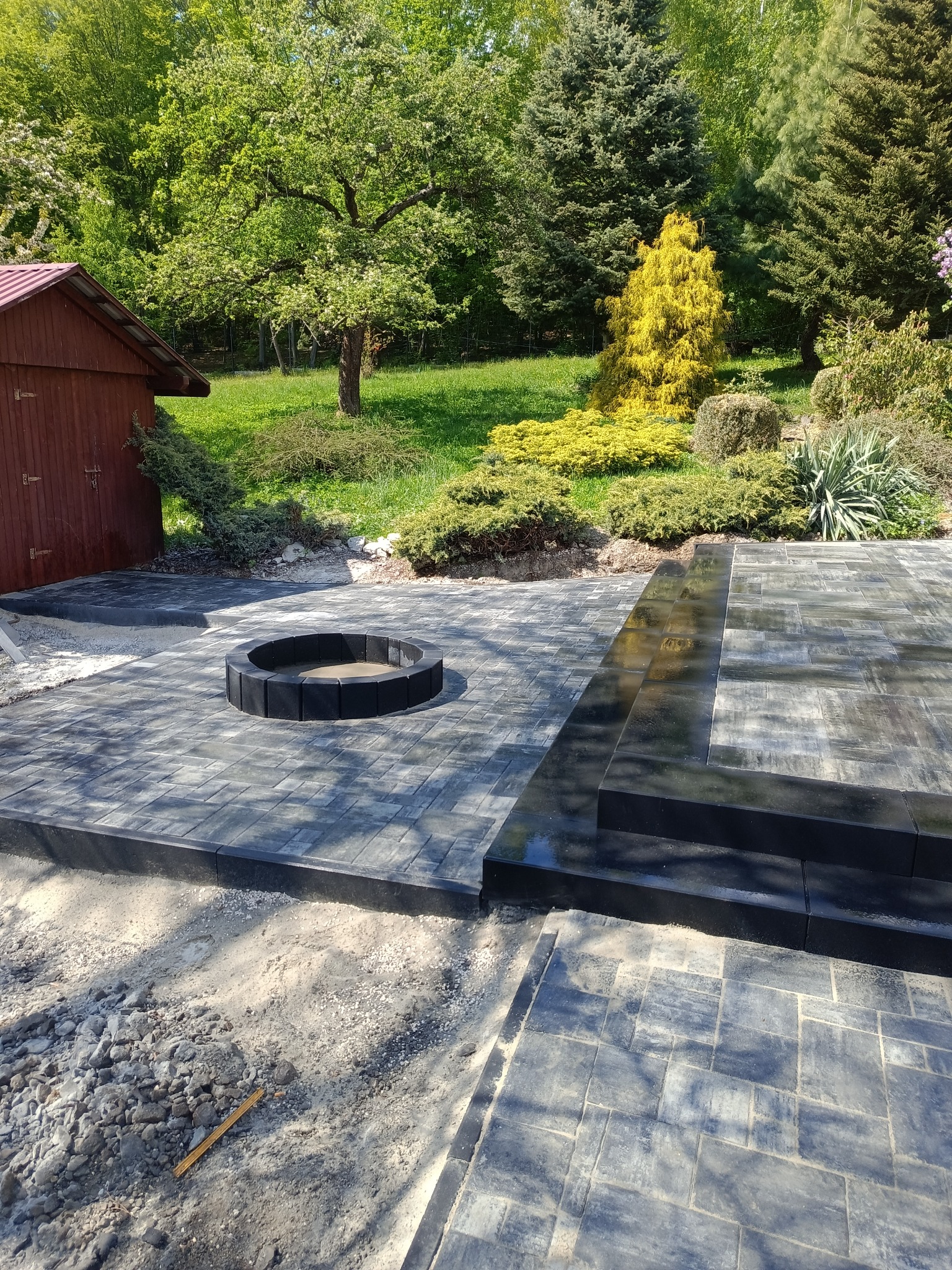 Wykończony taras z kostki brukowej w ogrodzie, z paleniskiem i schodami. Ujęcie z bliska na szare kostki i elementy paleniska na tle zieleni.
