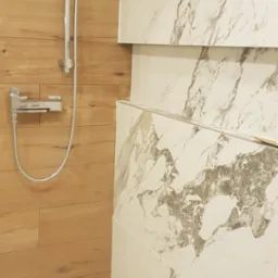 Nowoczesna kabina prysznicowa z drewnopodobnymi panelami i marmurowymi płytkami. Widoczna chromowana armatura i brodzik z folią ochronną.