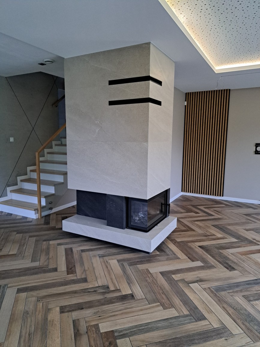 Nowoczesny salon z kominkiem obłożonym płytami imitującymi kamień, parkiet ułożony w jodełkę, drewniane lamele na ścianie i schody z białymi stopnicami.