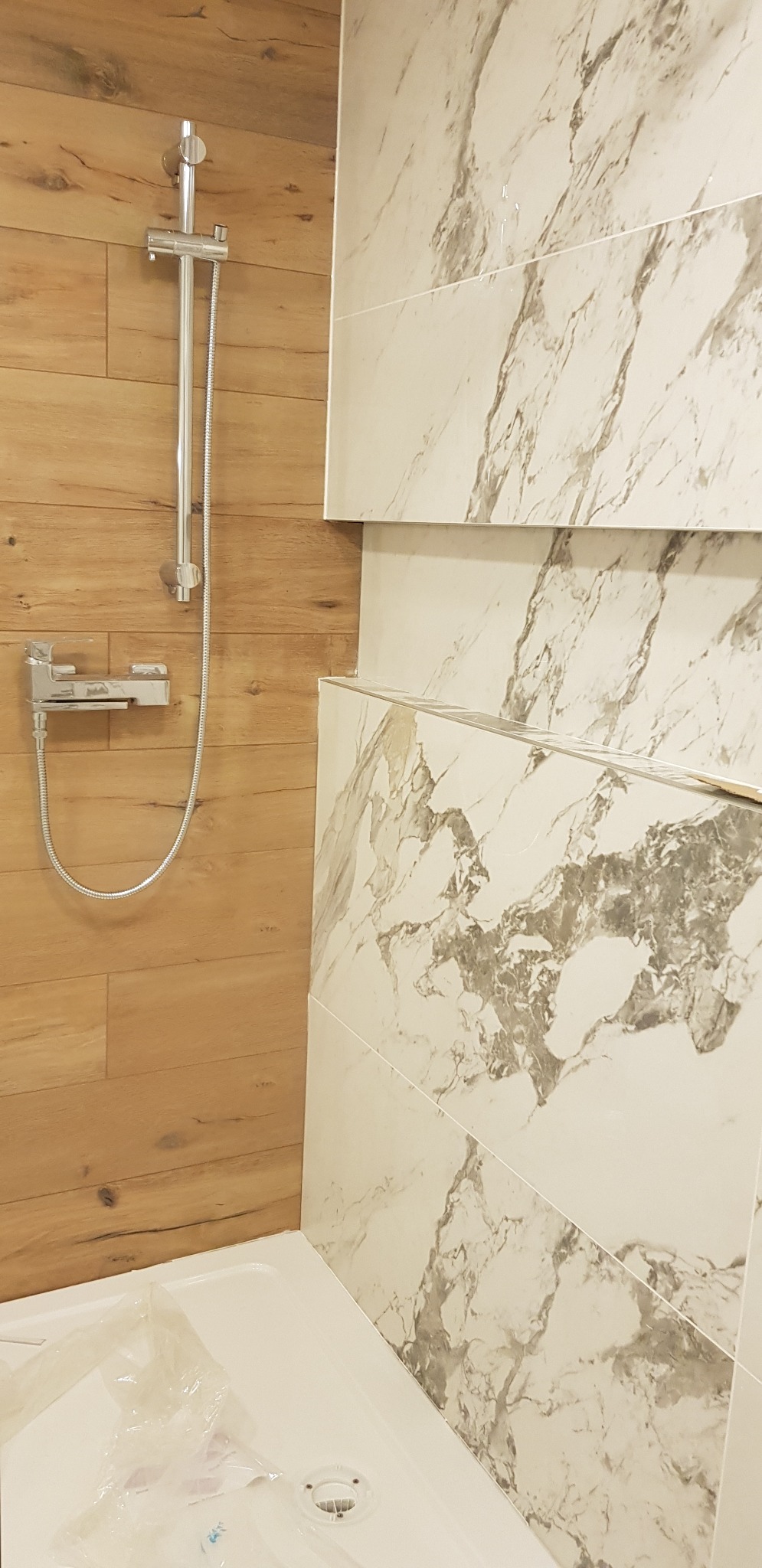 Nowoczesna kabina prysznicowa z drewnopodobnymi panelami i marmurowymi płytkami. Widoczna chromowana armatura i brodzik z folią ochronną.