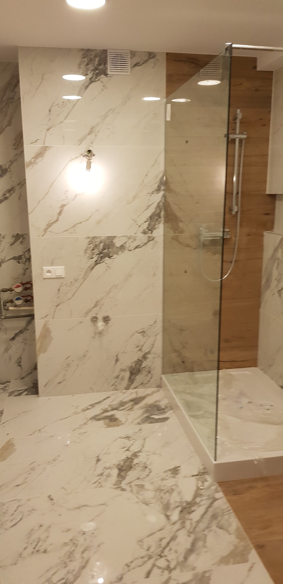 Nowoczesna kabina prysznicowa ze szklaną ścianką i marmurowymi płytkami w łazience. Widoczne elementy instalacji wodnej.