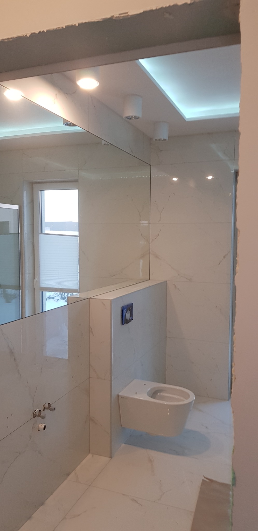 Biała łazienka z marmurową glazurą, podwieszana toaleta, lustro na ścianie odbija okno z żaluzjami. Oświetlenie sufitowe punktowe i ledowe.