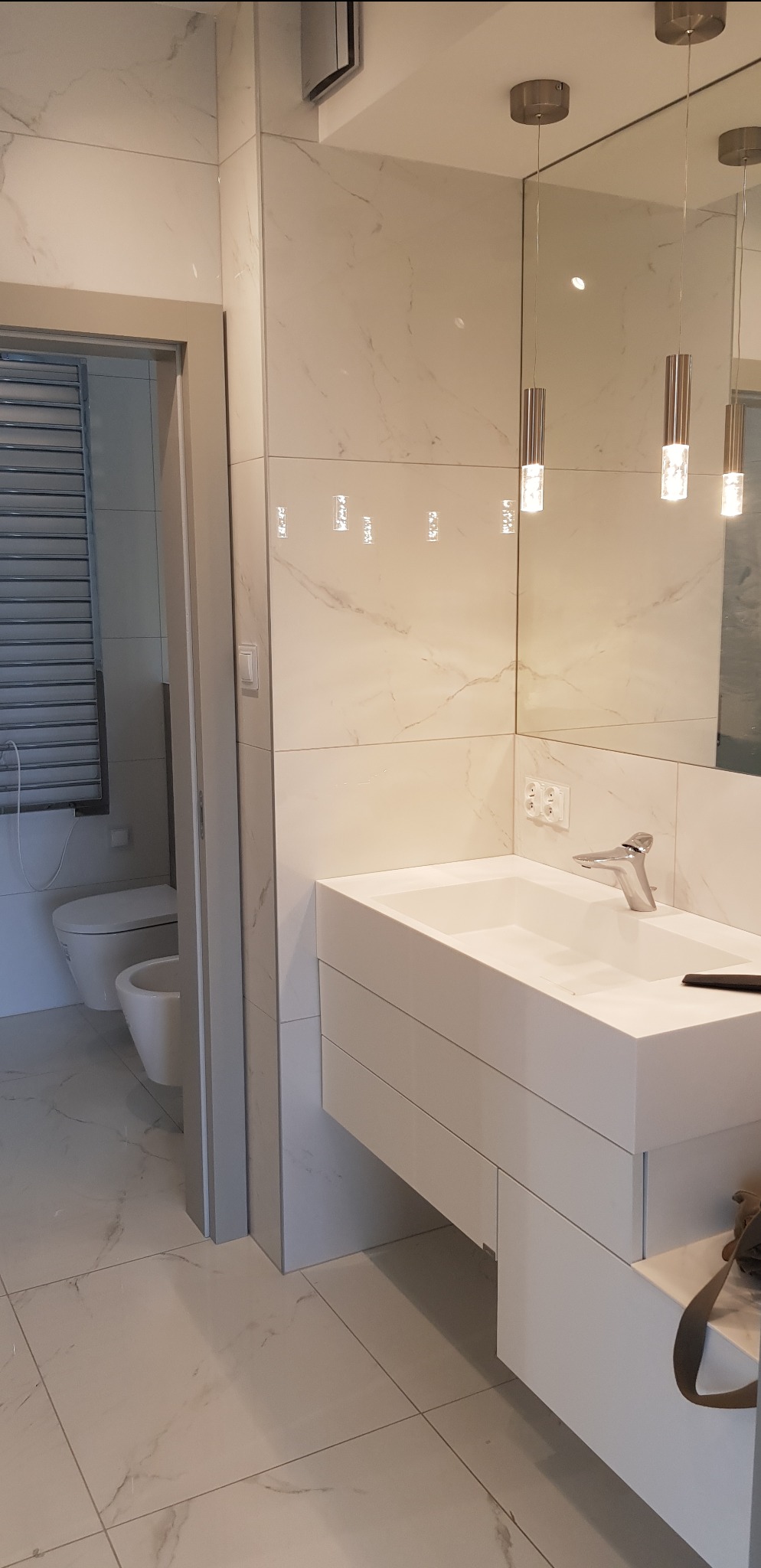 Nowoczesna łazienka z białą umywalką nablatową i marmurowymi płytkami. Widoczne oświetlenie punktowe i fragment toalety za otwartymi drzwiami. Minimalistyczny design.