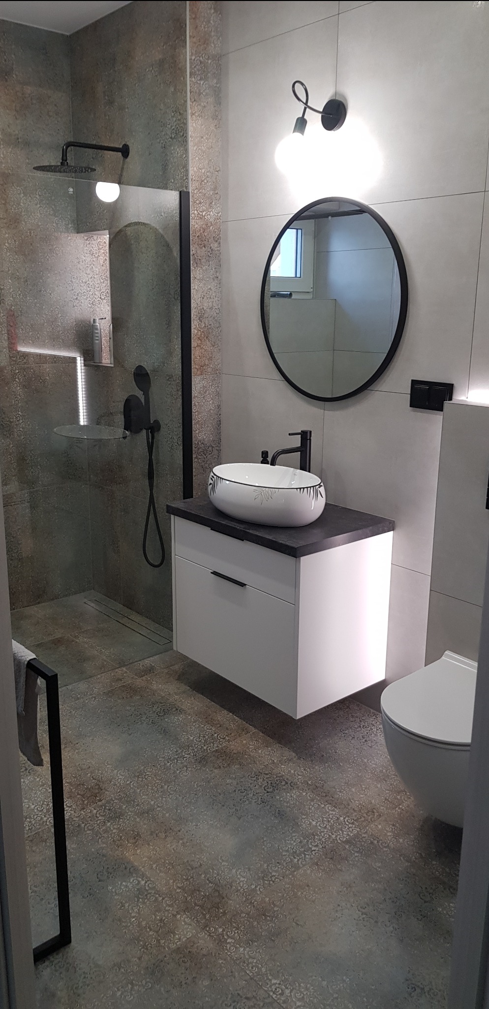 Nowoczesna łazienka z kabiną prysznicową typu walk-in, okrągłym lustrem z czarną ramą i umywalką z motywem liści. Całość utrzymana w szaro-białej kolorystyce.