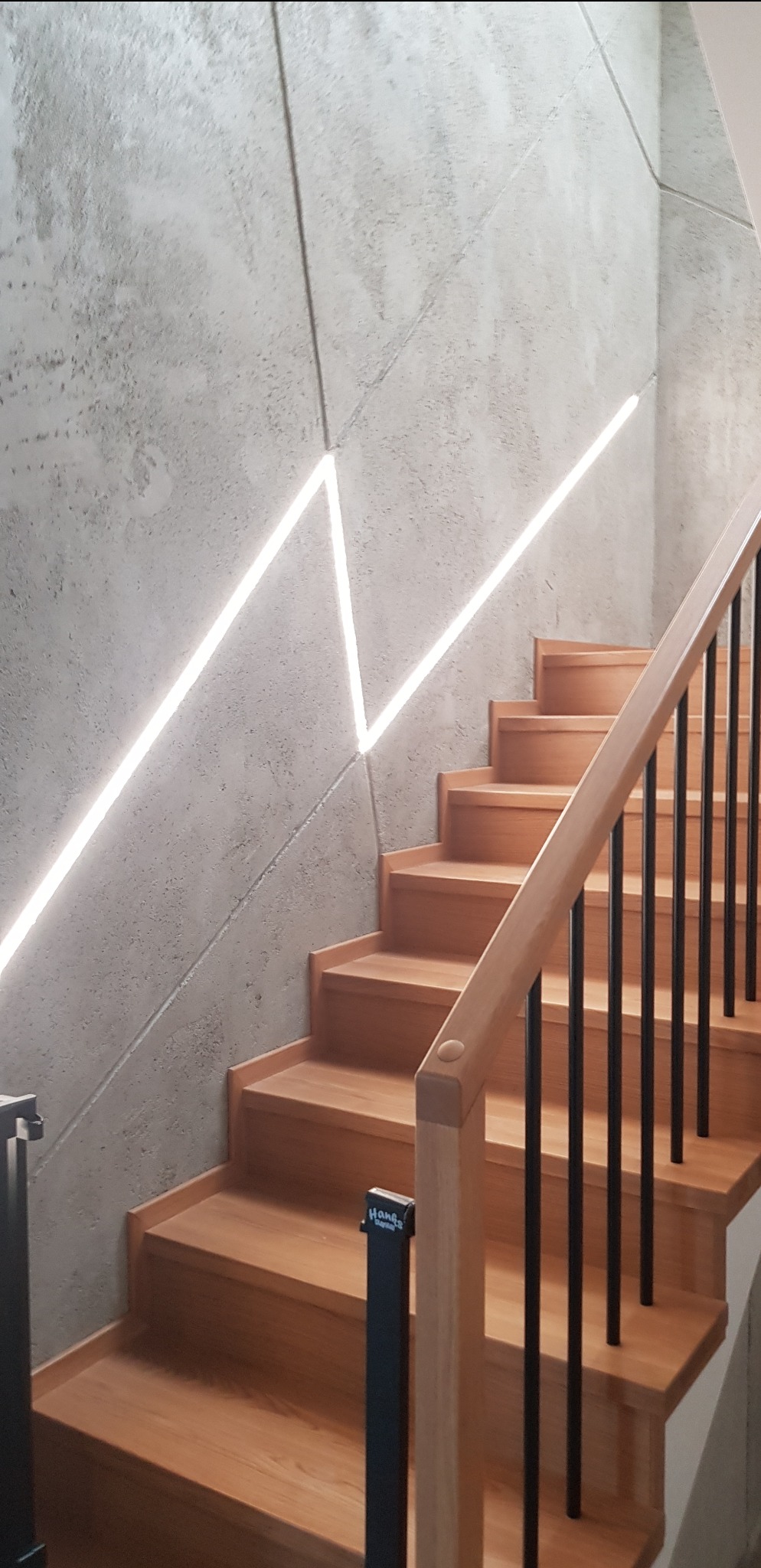 Nowoczesne schody z drewnianymi stopniami i metalową balustradą, oświetlone liniowym światłem LED wkomponowanym w surową, betonową ścianę.