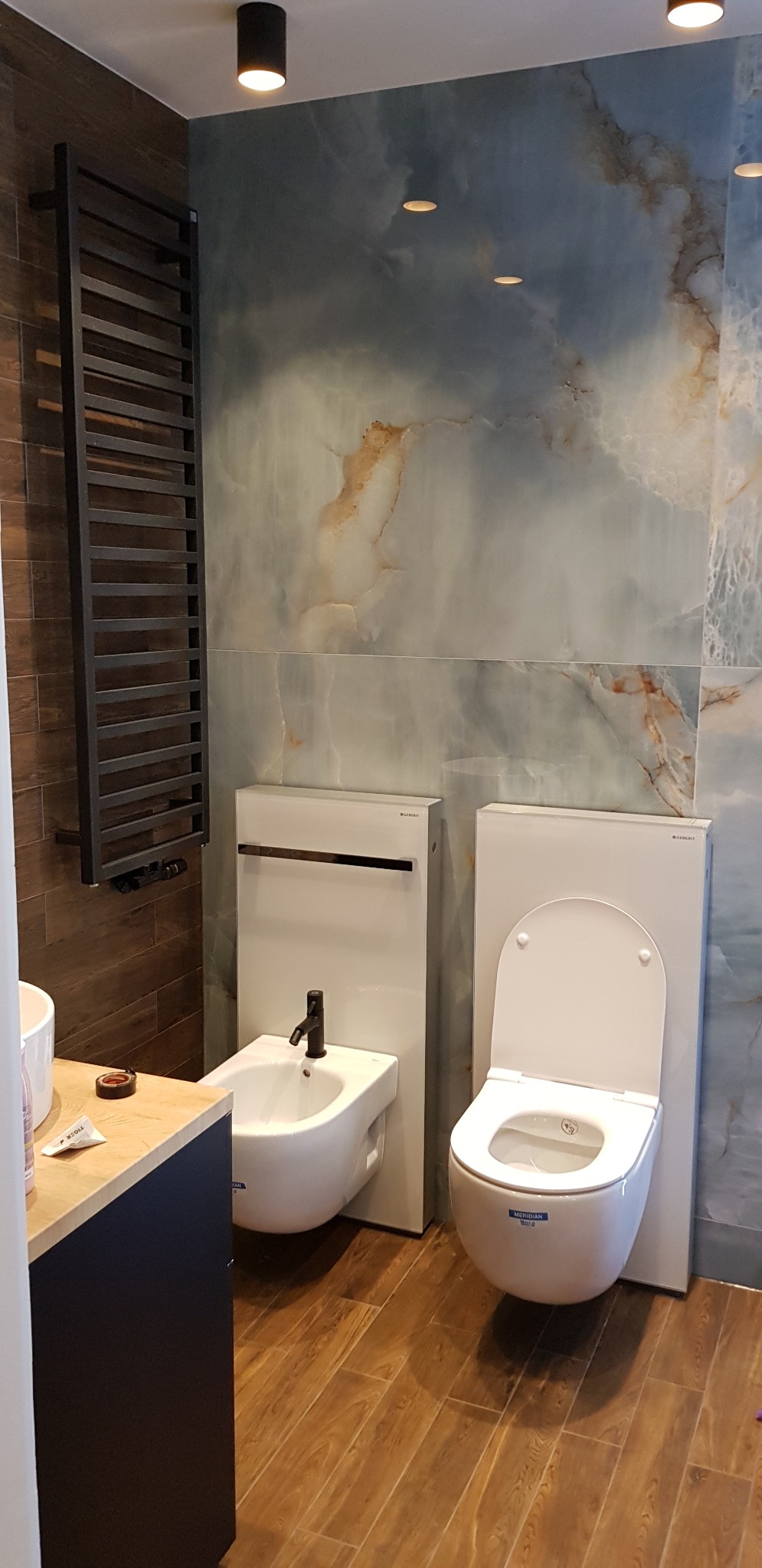 Wykończona łazienka w Łodzi: biała toaleta i bidet Geberit na tle marmurowej ściany, drewnopodobna podłoga i grzejnik drabinkowy. Nowoczesny design.