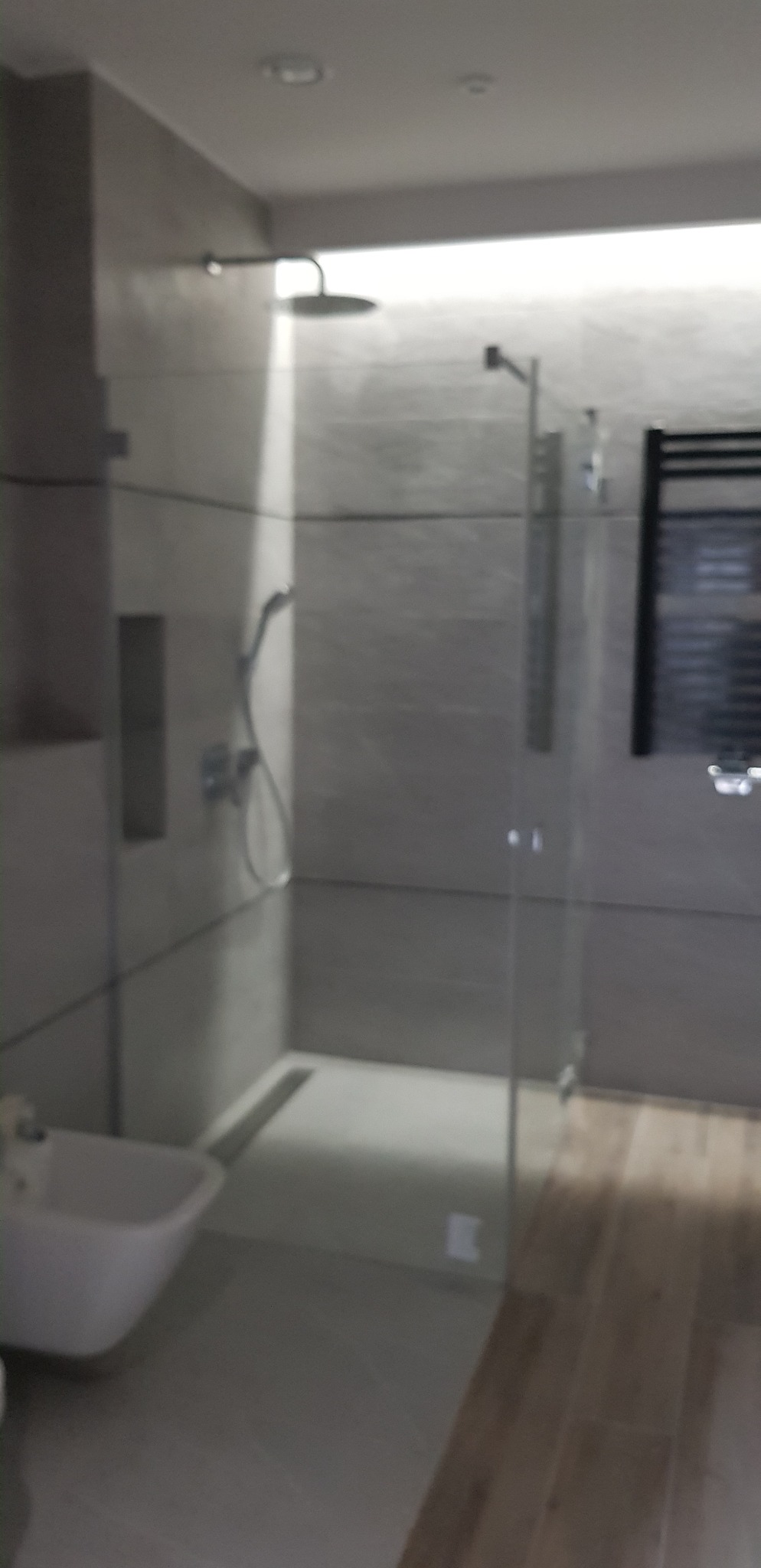 Nowoczesna łazienka z kabiną prysznicową typu walk-in, szarymi płytkami i drewnopodobną podłogą. Widoczny bidet i grzejnik drabinkowy.