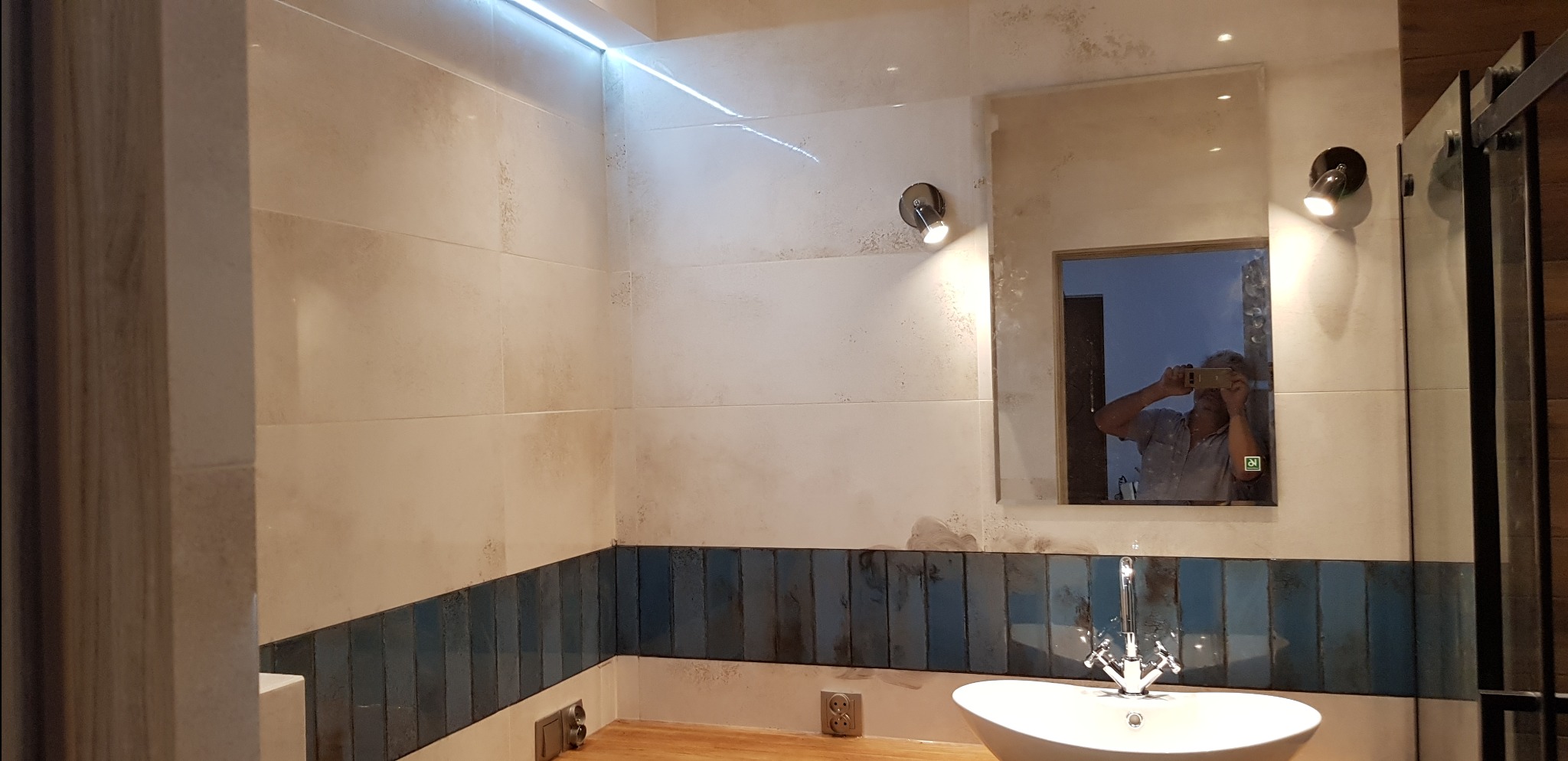 Wykończona łazienka z białymi i niebieskimi płytkami, owalną umywalką i lustrem z oświetleniem. W lustrze odbija się osoba robiąca zdjęcie.