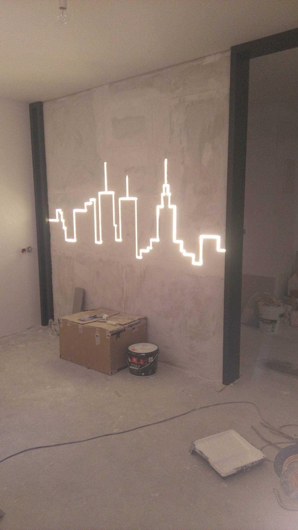 Surowa ściana z neonowym motywem miasta nad kartonem i puszką farby, widoczne elementy konstrukcyjne, podłoga w trakcie prac wykończeniowych.