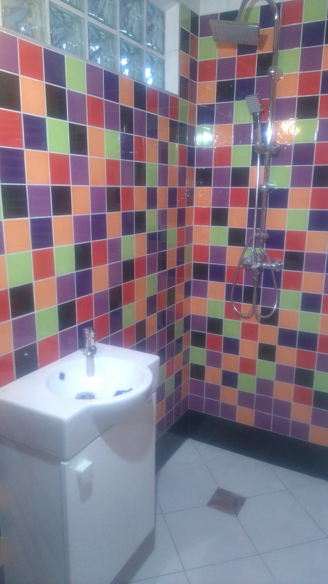 Kolorowa łazienka z umywalką i prysznicem. Ściany wyłożone są mozaiką z płytek w odcieniach fioletu, pomarańczy, czerwieni, zieleni i czerni. Biała umywalka z szafką.