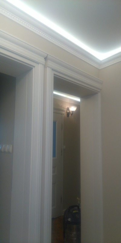 Białe, odnowione futryny drzwiowe z dekoracyjnym frezowaniem i oświetleniem LED w suficie. W tle widoczna lampa ścienna i odkurzacz.
