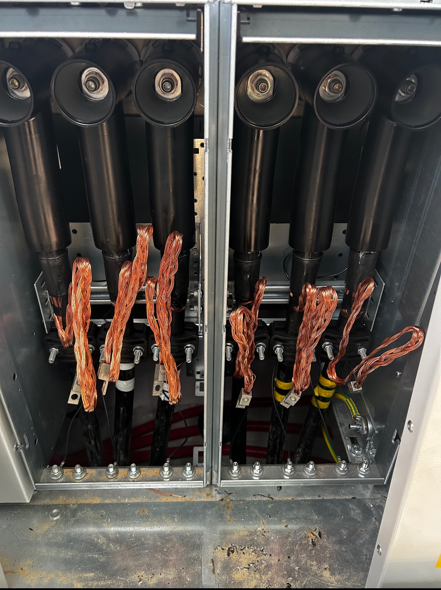 Wnętrze szafy elektrycznej z grubymi kablami zasilającymi i uziemieniem z plecionki miedzianej. Widoczne złącza i elementy montażowe w metalowej obudowie.