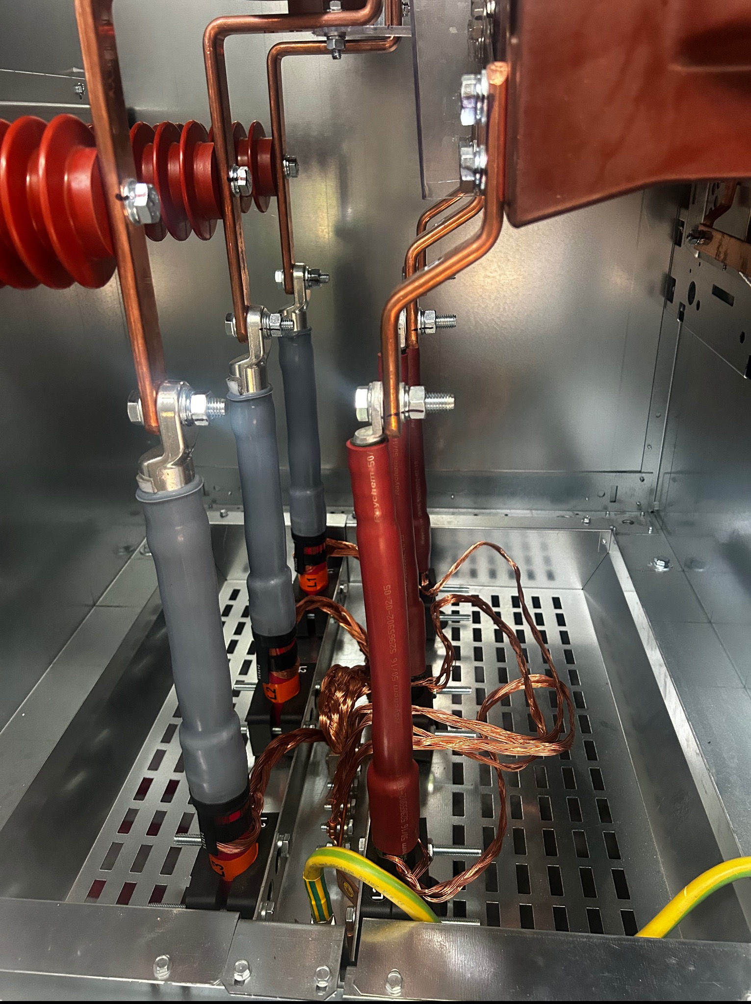 Wnętrze szafy elektrycznej z elementami instalacji: miedziane przewody, izolatory ceramiczne, kable w szarych i czerwonych osłonach, przewód uziemiający. Widok z góry.