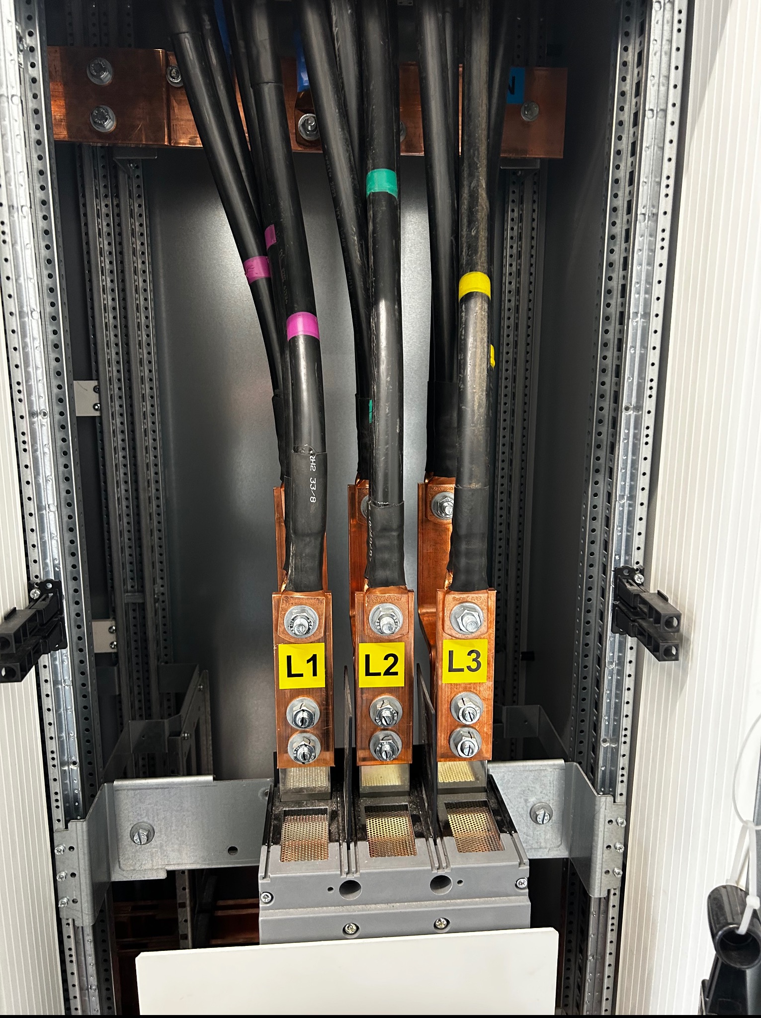 Kable zasilające L1, L2, L3, przymocowane do szyny w szafie elektrycznej. Widoczne kolorowe oznaczenia kabli i elementy montażowe.
