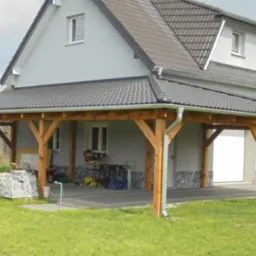 Drewniana konstrukcja zadaszenia nad podjazdem, z szarym, dachówkopodobnym pokryciem, przylegająca do budynku z szarą elewacją i białymi oknami; widoczne rynny, narzędzia i materiały budowlane.