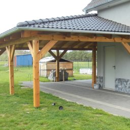 DACH-BUD COMPLEX Jerzy Orankiewicz - Drewniana wiata garażowa z ciemną dachówką ceramiczną przylegająca do jasnego budynku, z widocznymi rynnami i elementami więźby dachowej, na trawniku.