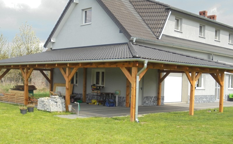 Drewniana konstrukcja zadaszenia nad podjazdem, z szarym, dachówkopodobnym pokryciem, przylegająca do budynku z szarą elewacją i białymi oknami; widoczne rynny, narzędzia i materiały budowlane.