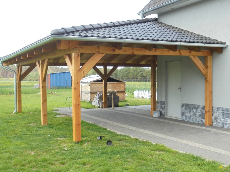 Drewniana wiata garażowa z ciemną dachówką ceramiczną przylegająca do jasnego budynku, z widocznymi rynnami i elementami więźby dachowej, na trawniku.