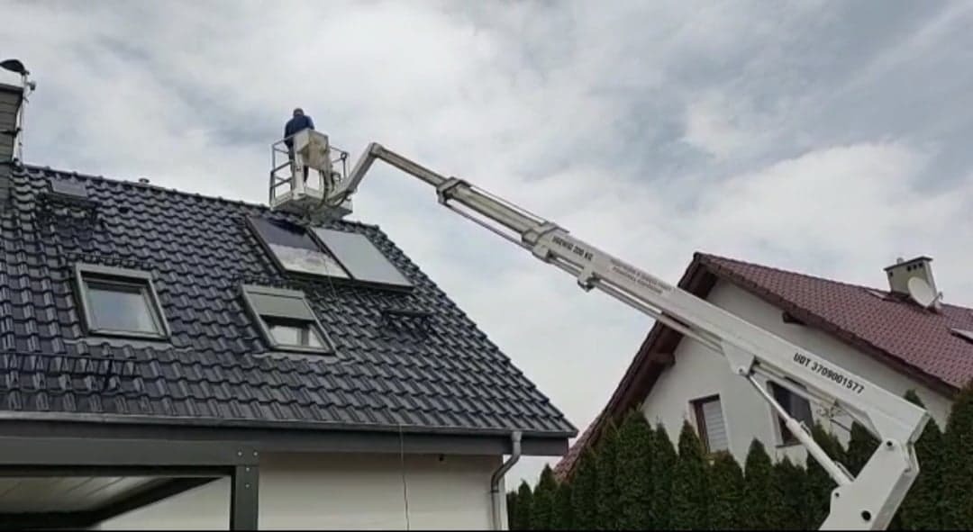 Pracownik na podnośniku koszowym montuje panele słoneczne na dachu z ciemnej dachówki, widoczne okna dachowe i rynna, w tle budynek z czerwoną dachówką.