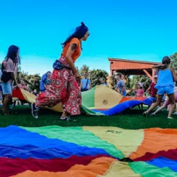 Animator prowadzi zabawy z kolorową chustą animacyjną na trawie, w tle grupa dzieci i rodziców, błękitne niebo.