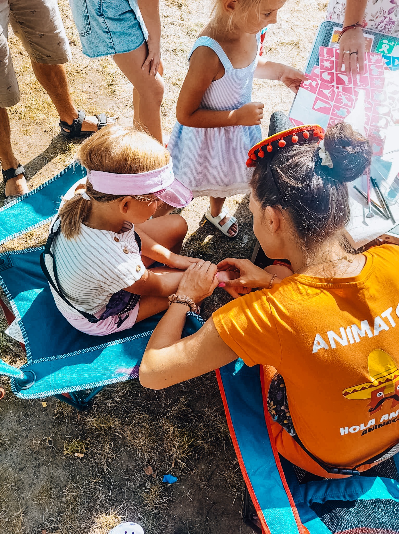 Animator maluje brokatowy tatuaż na dłoni dziewczynki siedzącej na turkusowym krzesełku; na stoliku szablony tatuaży i akcesoria.
