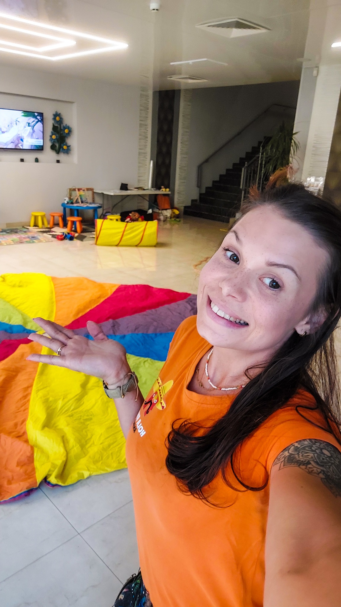 Uśmiechnięta kobieta w pomarańczowej koszulce prezentuje kolorową chustę animacyjną na tle sali zabaw z tunelem i stolikami dla dzieci.