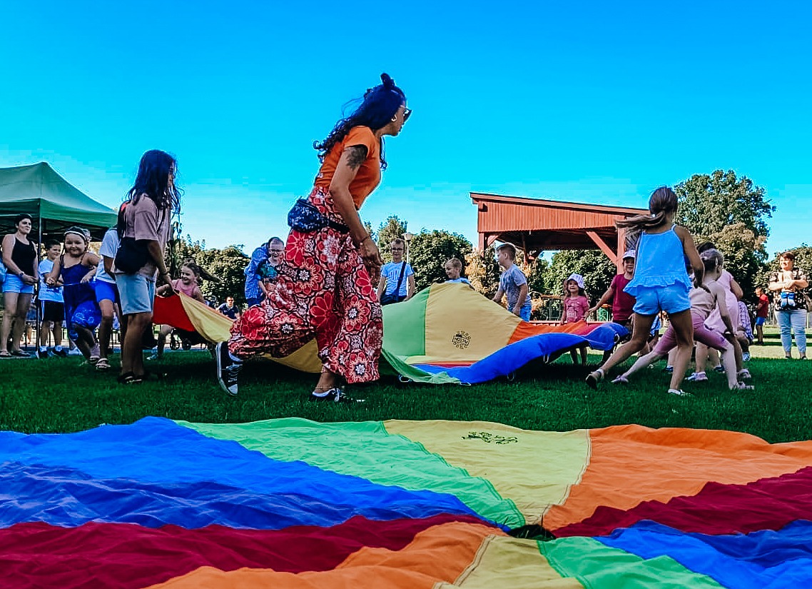 Animator prowadzi zabawy z kolorową chustą animacyjną na trawie, w tle grupa dzieci i rodziców, błękitne niebo.