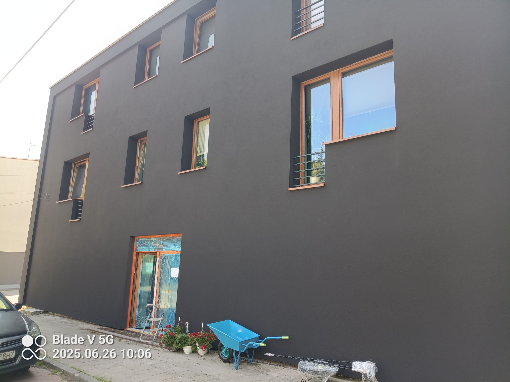 Elewacja budynku po renowacji w Ogrodzieńcu, pomalowana na ciemny kolor. Widoczne okna z drewnianą ramą, zabezpieczone wejście i taczka.