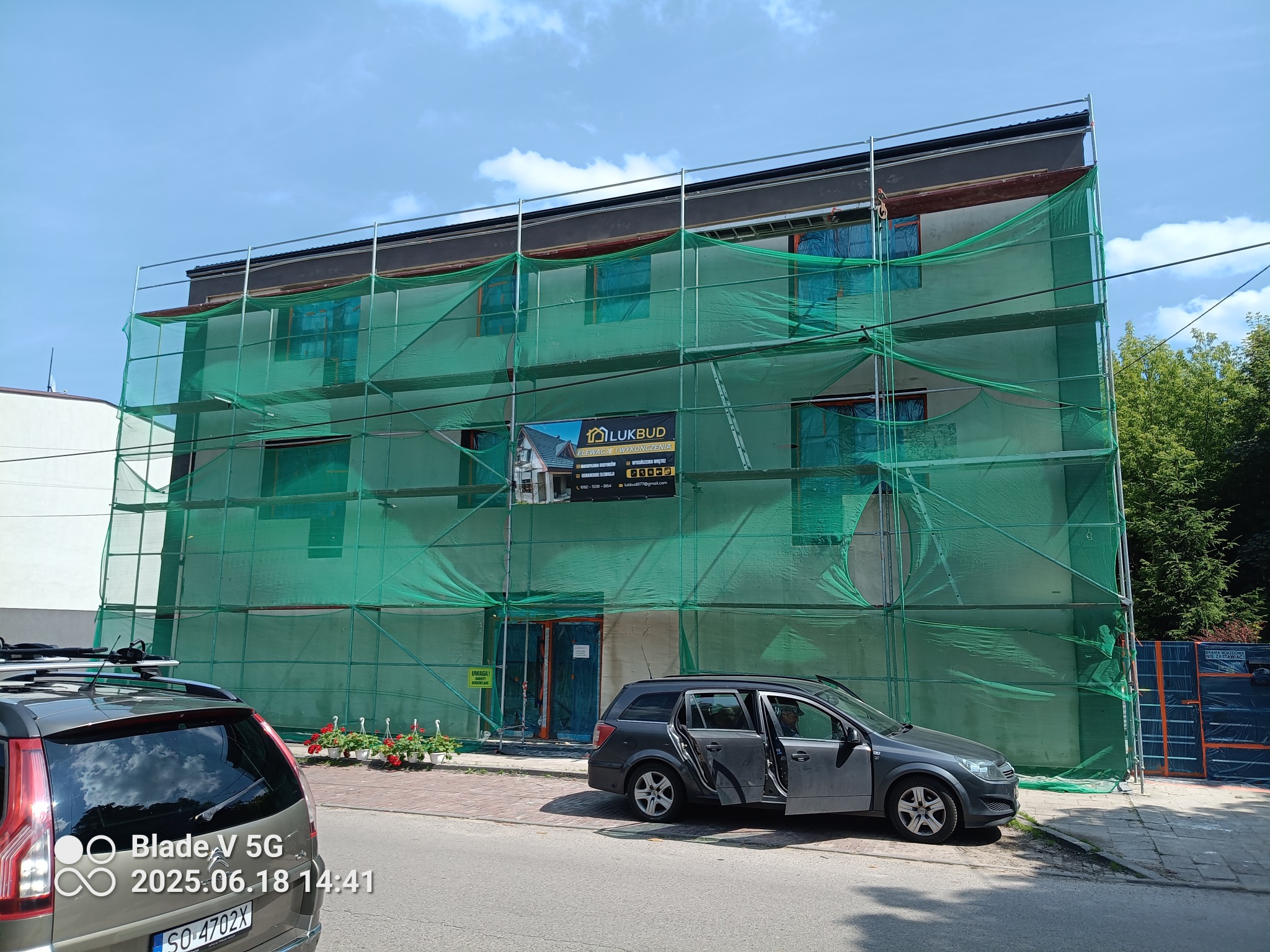 Budynek w trakcie prac elewacyjnych, osłonięty zieloną siatką na rusztowaniach. Widoczny baner firmy budowlanej LUKBUD. Na pierwszym planie zaparkowany samochód.