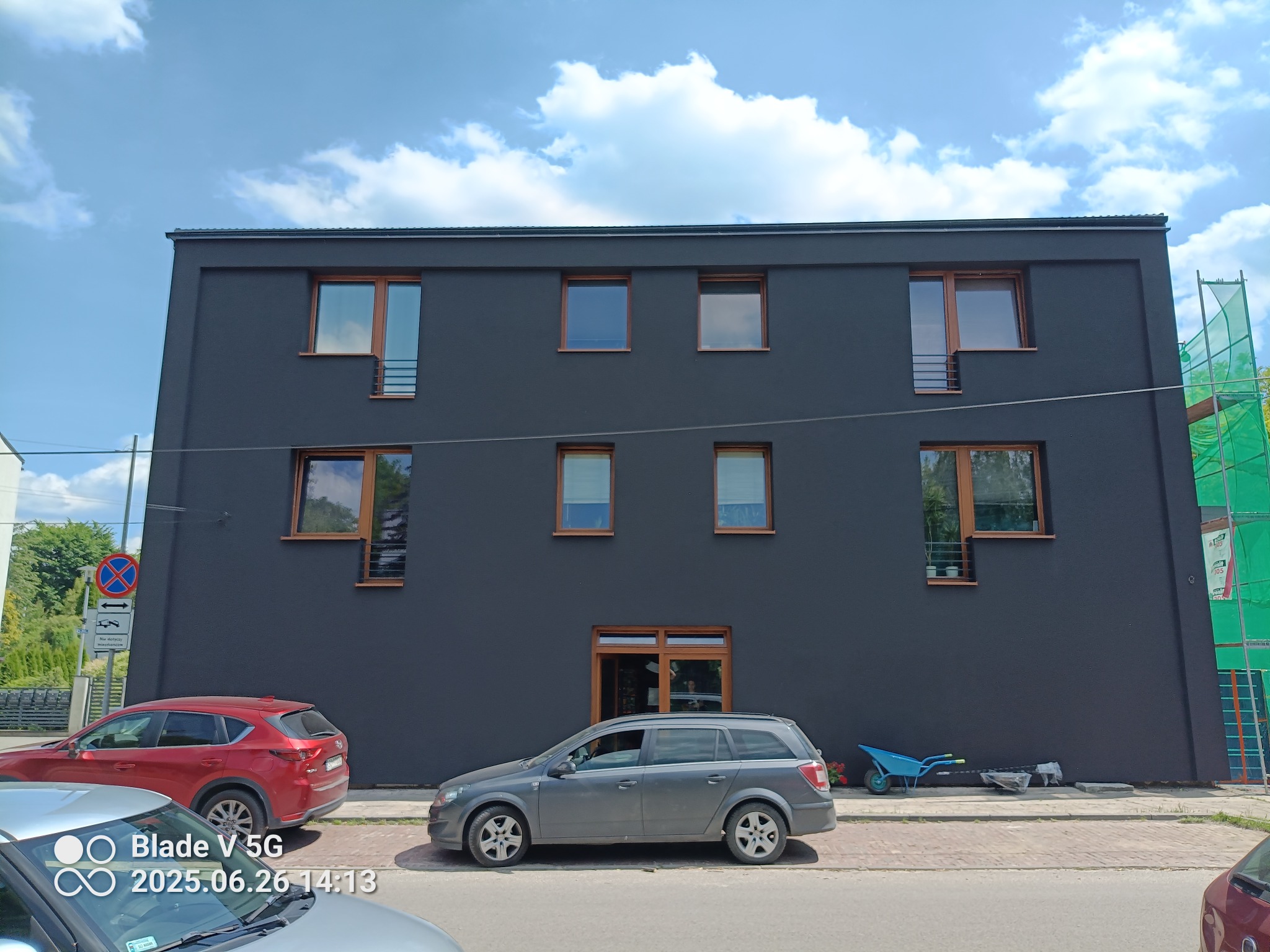Nowoczesny, dwupiętrowy budynek z ciemną elewacją i drewnianymi oknami. Na chodniku zaparkowane samochody i taczka. Częściowo widoczne rusztowanie z boku budynku.
