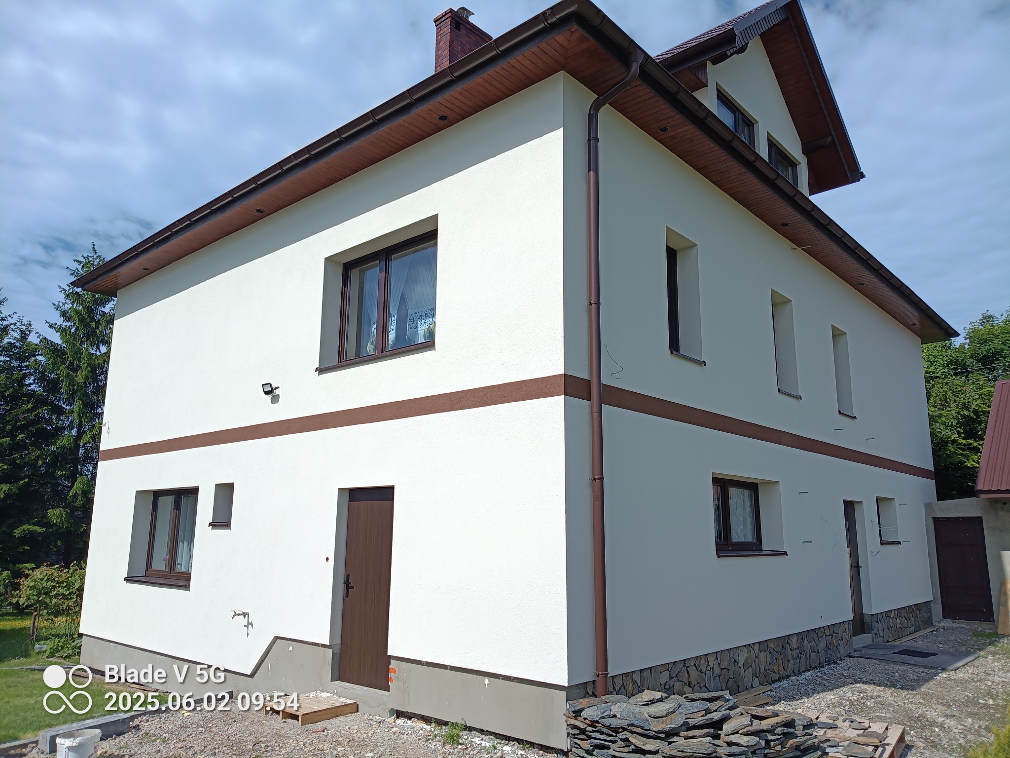 Dwukondygnacyjny dom z jasną elewacją, brązowymi akcentami i kamienną podmurówką, uchwycony pod kątem z widocznym fragmentem dachu i otoczenia.