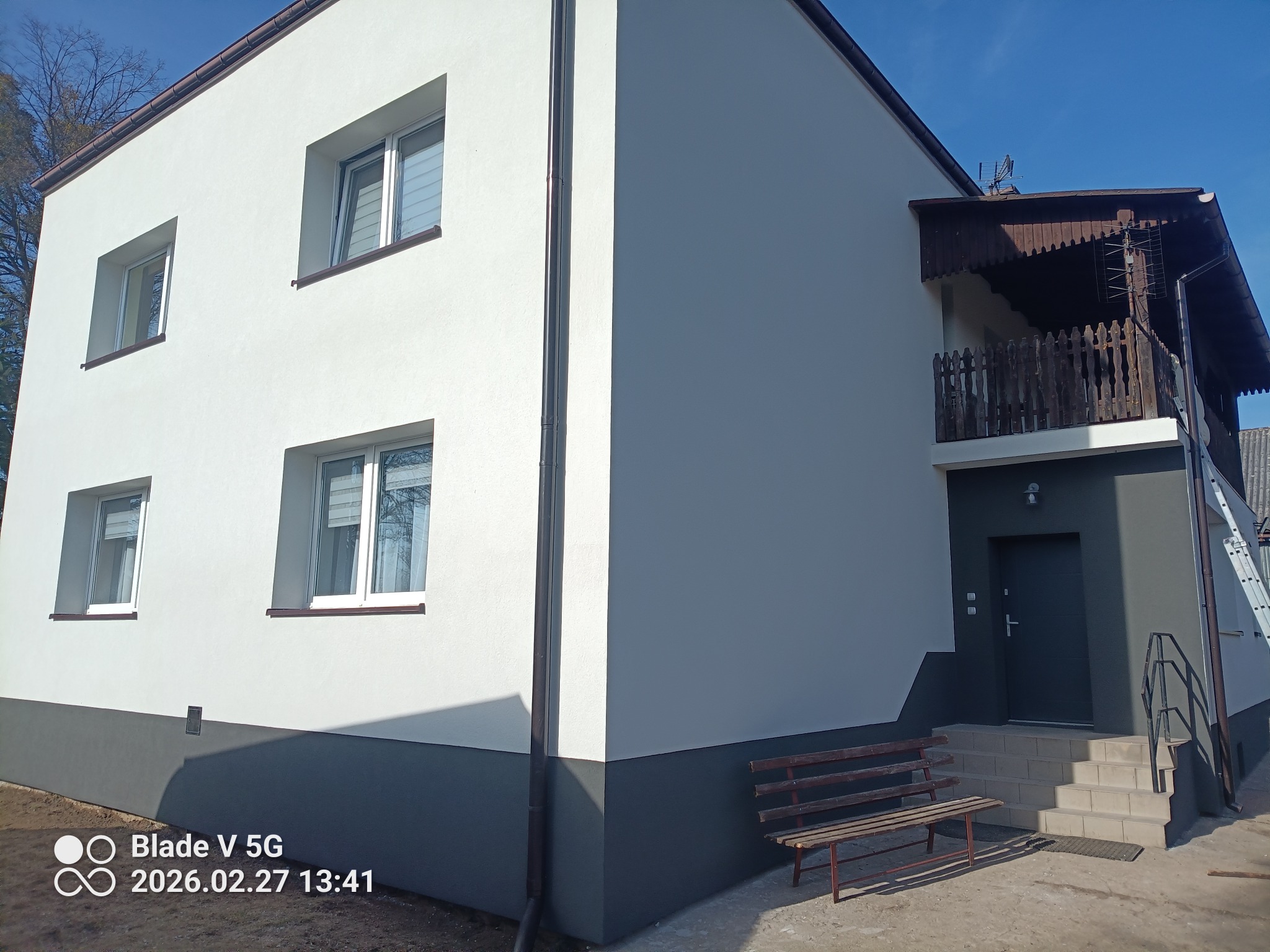 Odnowiony dom z białą elewacją i szarym cokołem, widoczny z boku, z drewnianą ławeczką przed wejściem i balkonem z drewnianą balustradą.