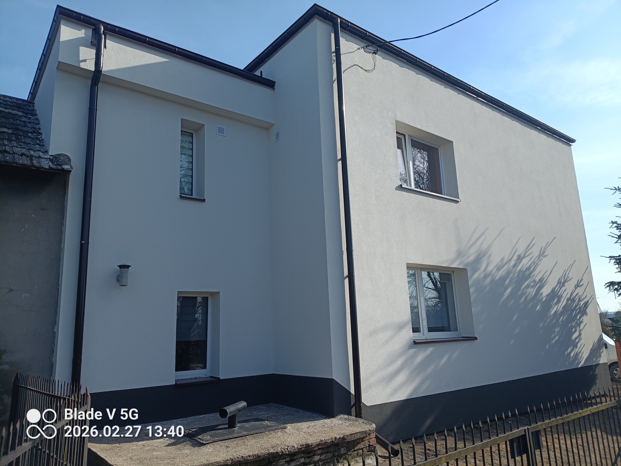 Dwukondygnacyjny budynek po renowacji elewacji, z widocznymi oknami i rynnami. Świeżo pomalowana fasada w jasnym odcieniu, z ciemniejszym cokołem. Ujęcie z poziomu gruntu.