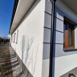 Dariusz Lewandowski - Świeżo pomalowana elewacja domu jednorodzinnego w odcieniach szarości i bieli, z widoczną rynną, oknem z drewnianą ramą i cieniem drzewa na ścianie.